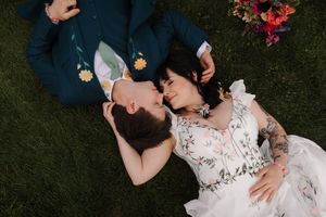 Aerial view of bride and groom lying on green grass, sharing a romantic moment in wedding attire.