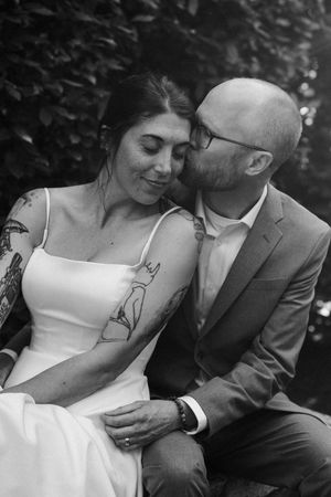 Black and white wedding photo of groom kissing tattooed bride on cheek while sitting together outdoors.