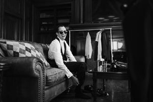 A dramatic black and white photo of a person in a suit and glasses sitting in a dimly lit room.