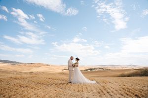 A couple stands together in a golden wheat field under a bright blue sky with white puffy clouds.