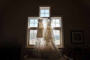 A wedding dress hangs elegantly in front of a sunlit window, creating a romantic silhouette against white shutters.