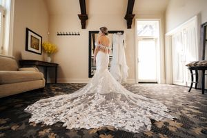 A stunning white lace wedding dress with a dramatic cathedral train spreads across a hardwood floor in natural light.