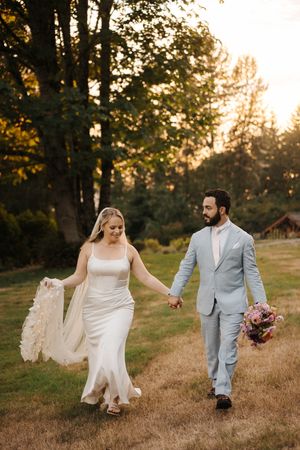 A couple in wedding attire walks together through a golden sunlit field during sunset.