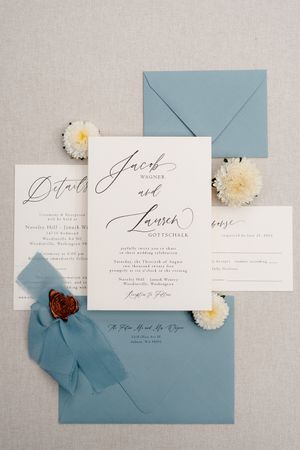 Elegant wedding invitation suite featuring calligraphy on white cards with dusty blue envelopes and white floral accents.