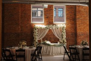Elegant wedding reception setup with floral arch and white drapes against exposed brick wall with ambient lighting.