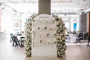 A white floral wedding ceremony arch stands in an industrial warehouse space with concrete pillars and exposed ceiling.