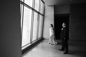 A couple in formal wedding attire stands in a minimalist hallway with large windows creating dramatic lighting.