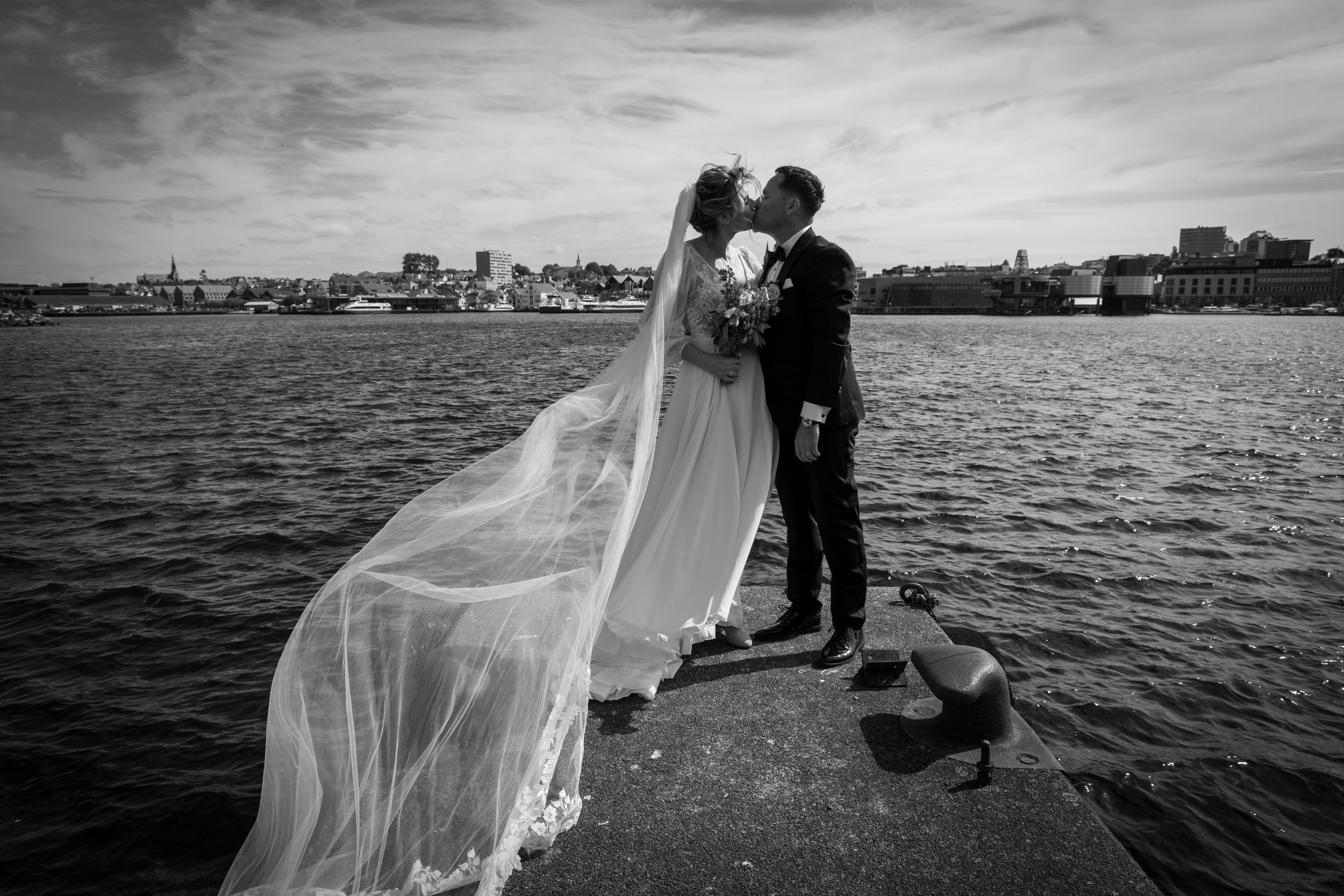 Black and white romantic wedding photo of couple kissing by the waterfront with a long flowing veil.