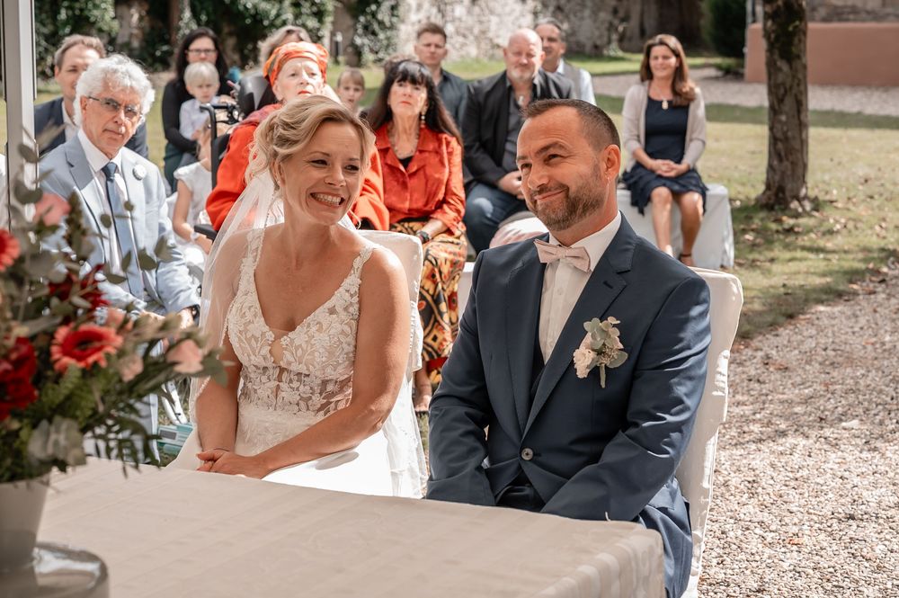 Wedding guests sit on wooden chairs during an outdoor ceremony at a rustic venue with gravel paths.