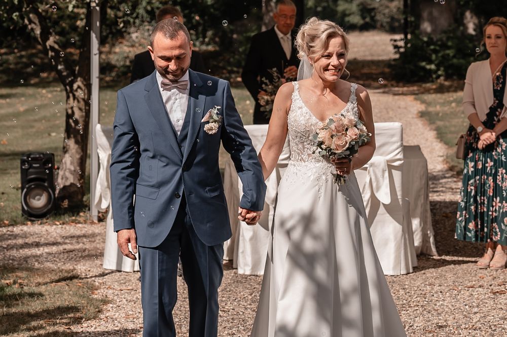 A couple walks together outdoors on their wedding day, wearing a blue suit and white wedding gown in summer sunlight.