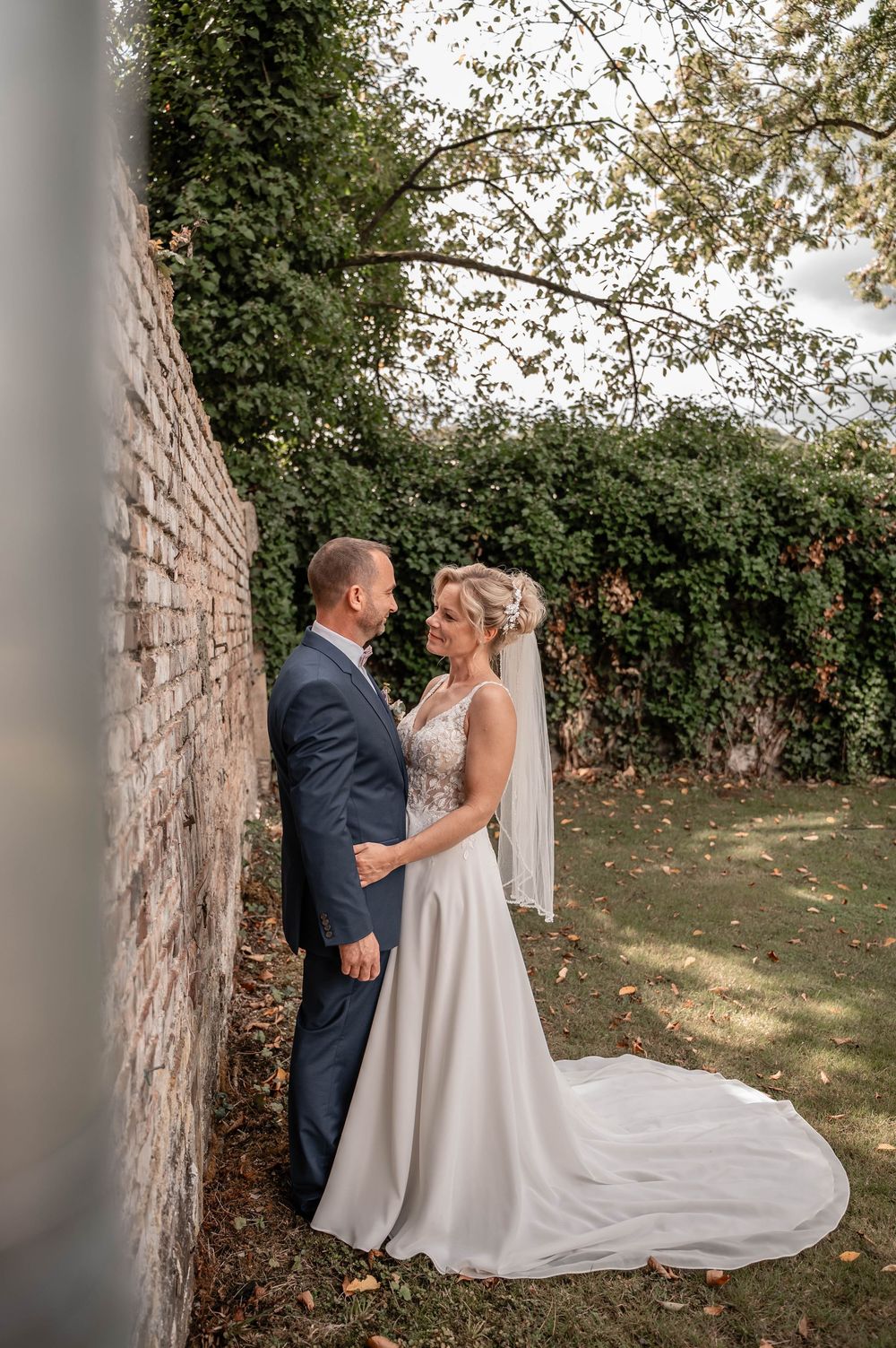 A couple in formal wedding attire embrace against a stone wall with lush greenery and trees in the background.