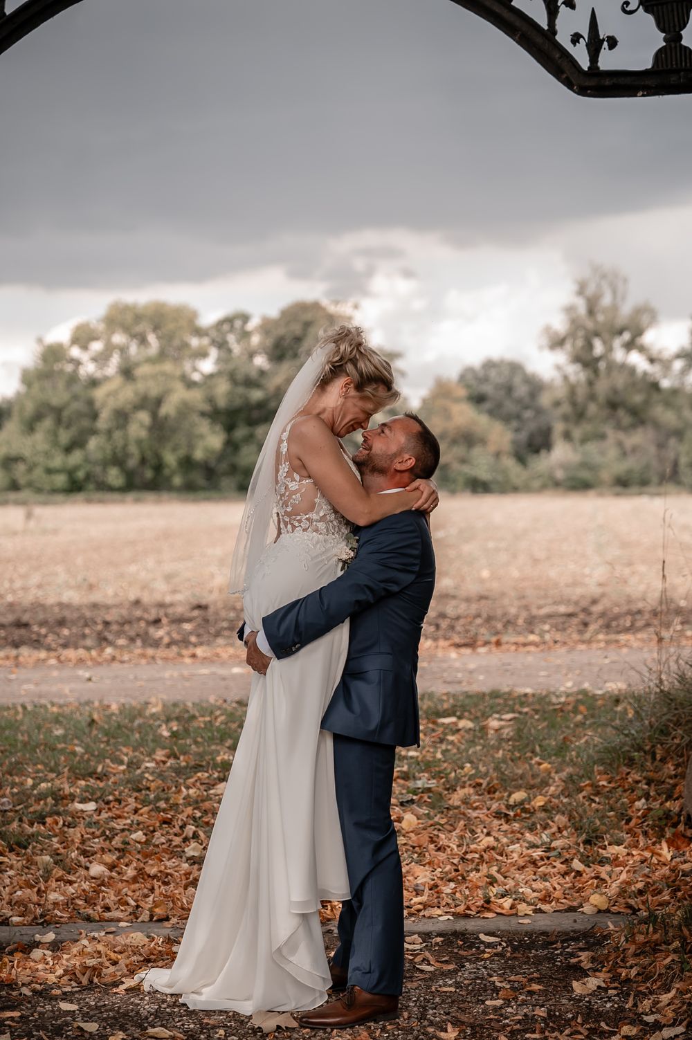 A couple embraces in an autumn field with fallen leaves and trees in the background on their wedding day.