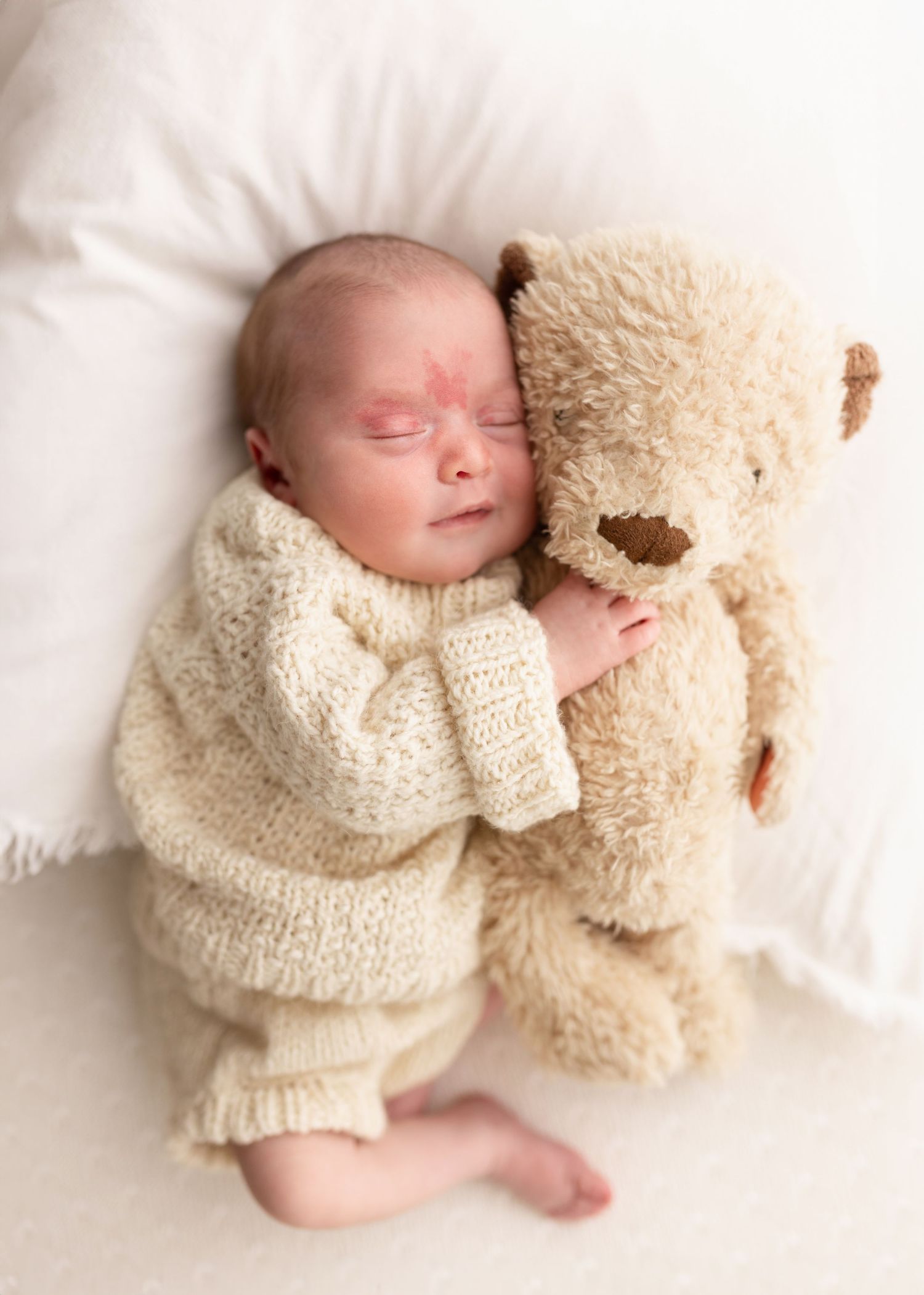 Sleeping baby in cream knit sweater cuddling with large beige teddy bear on white bedding.