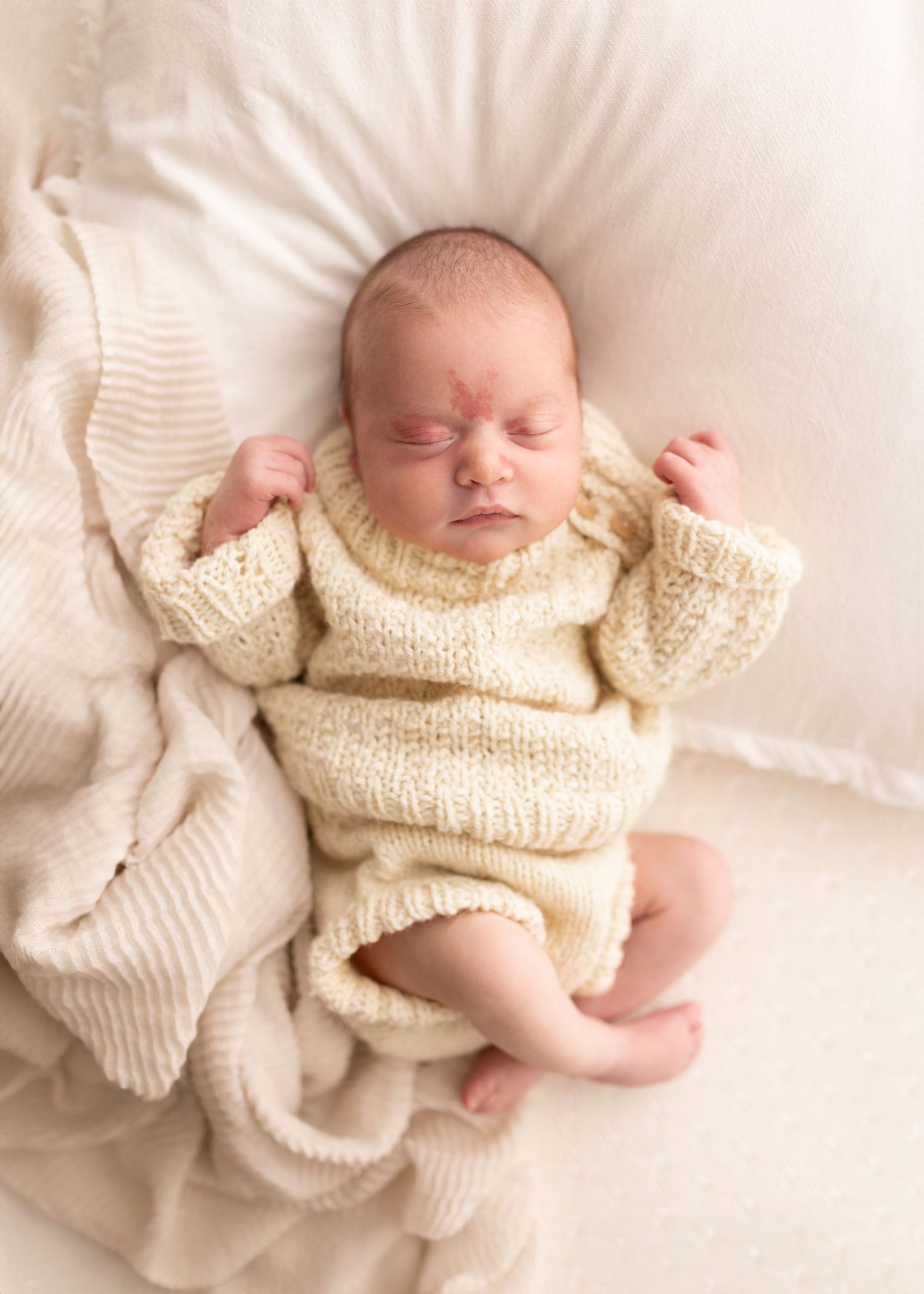 A sleeping newborn baby wearing a cream knit sweater lies peacefully on white bedding.