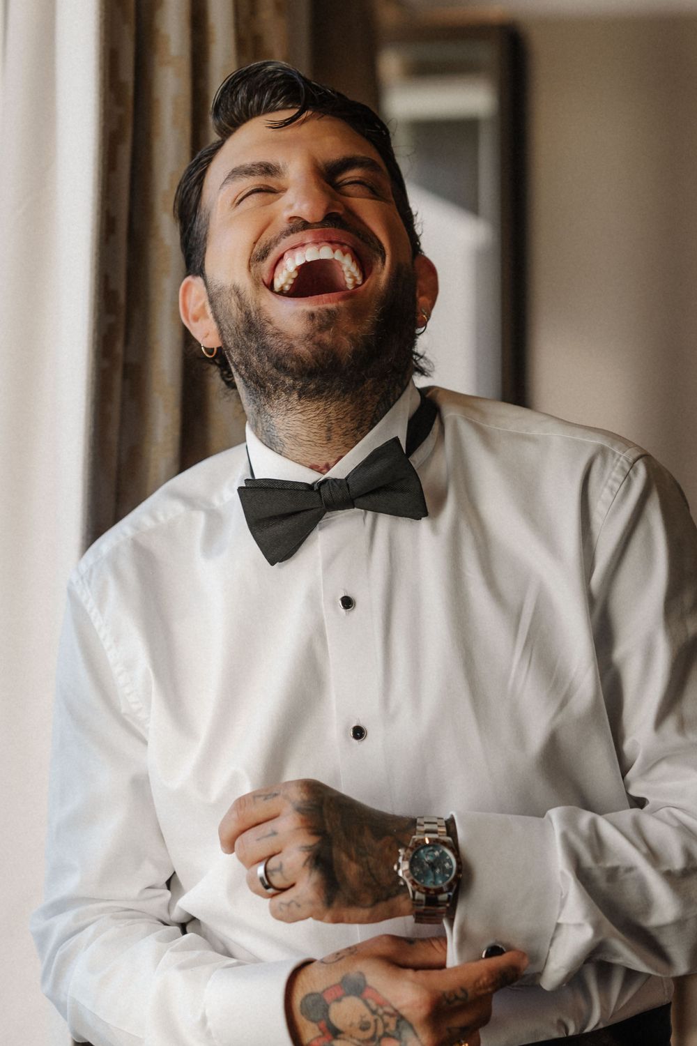 A groom in a white dress shirt and bow tie laughs joyfully during wedding preparations.