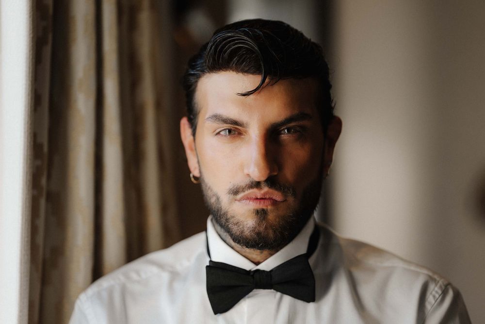 A person in a black suit and bow tie adjusts their collar while standing near curtained windows.