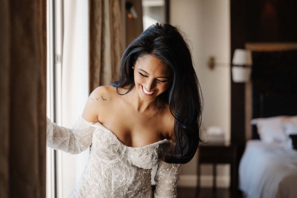 Elegant off-shoulder lace wedding dress being fitted in a sunlit bridal preparation room.