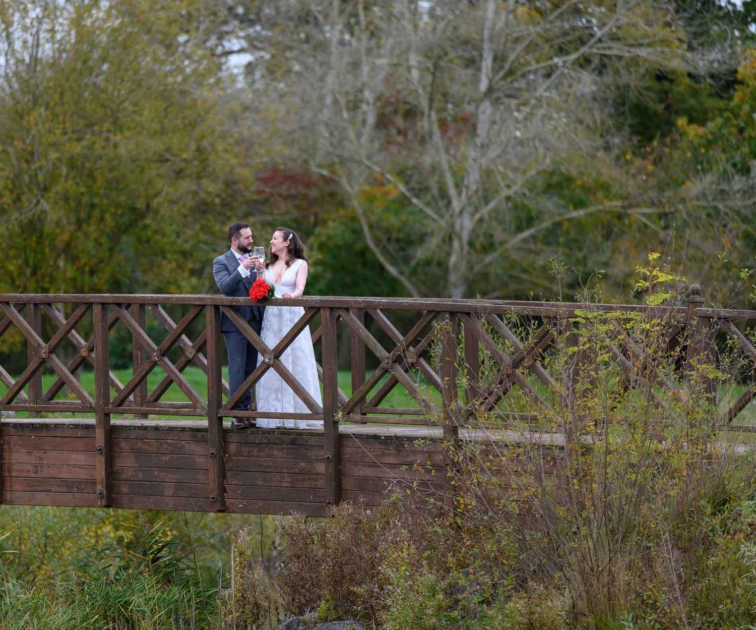 Crows Nest Barton wedding photographer captures a bride and groom sharing glasses of champagne on a wooden bridge.