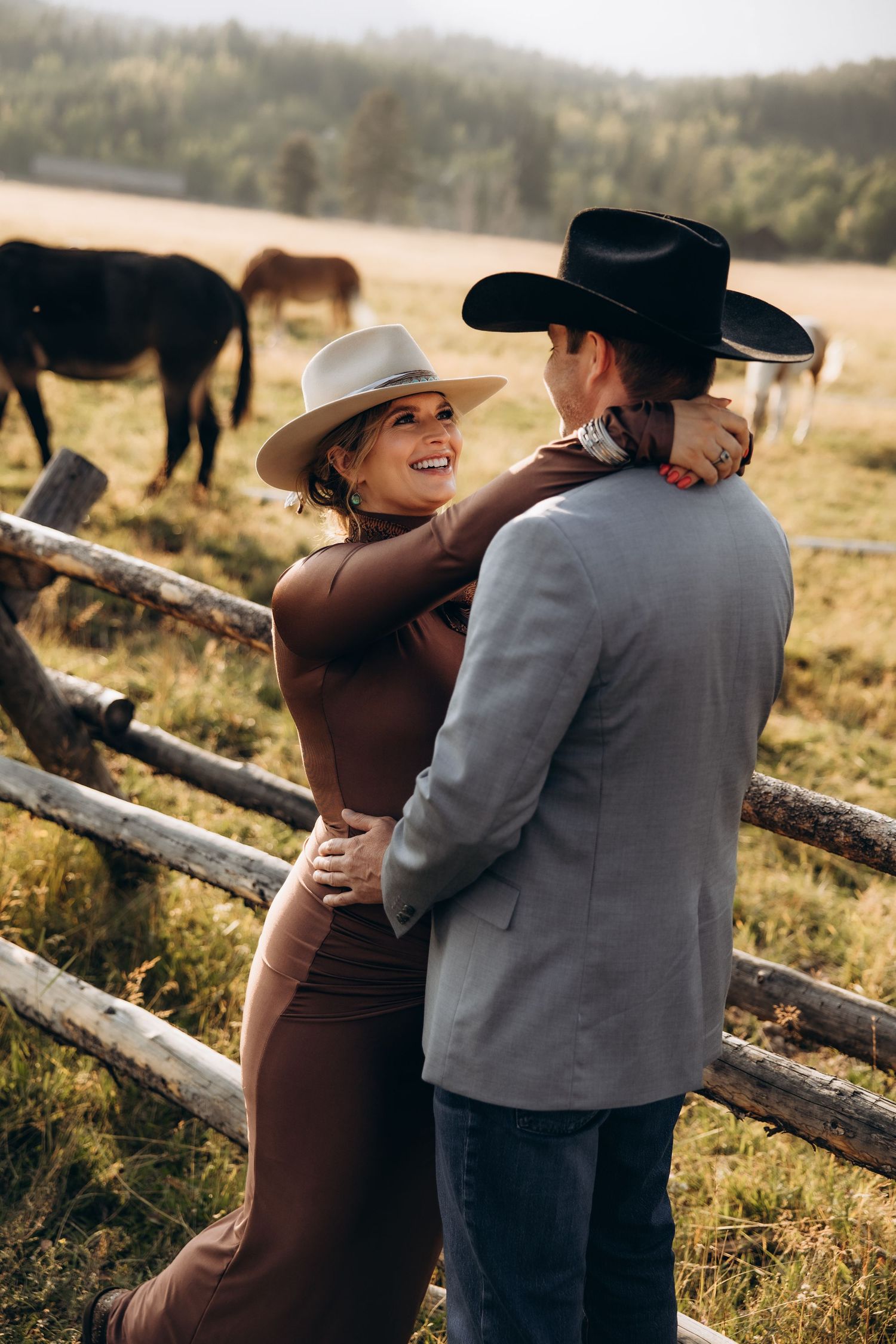 Couple shares intimate moment against rustic wooden fence on golden field with horses grazing in background.