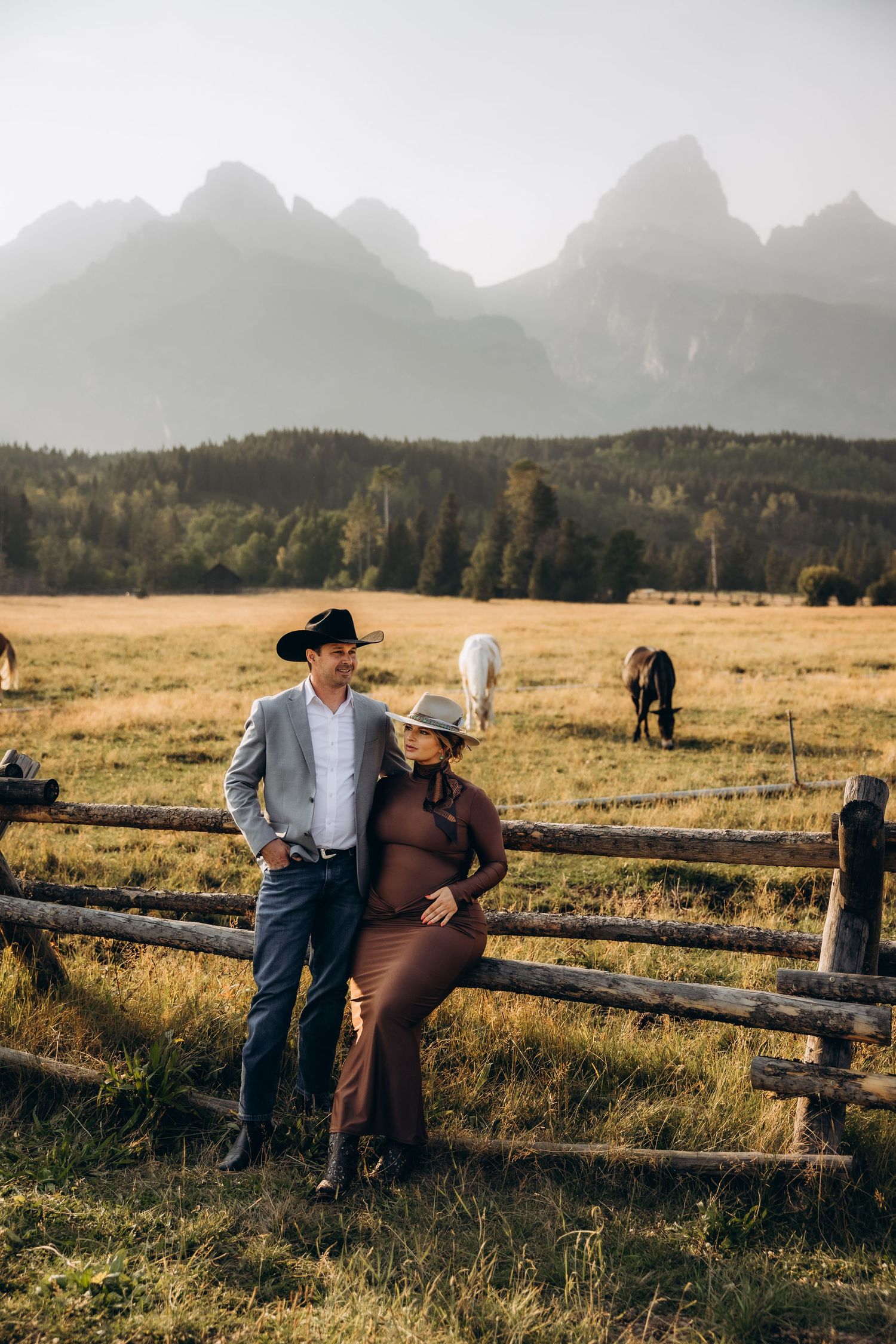 Couple sits together on rustic wooden fence with majestic Grand Teton mountains rising in the misty background at sunset.