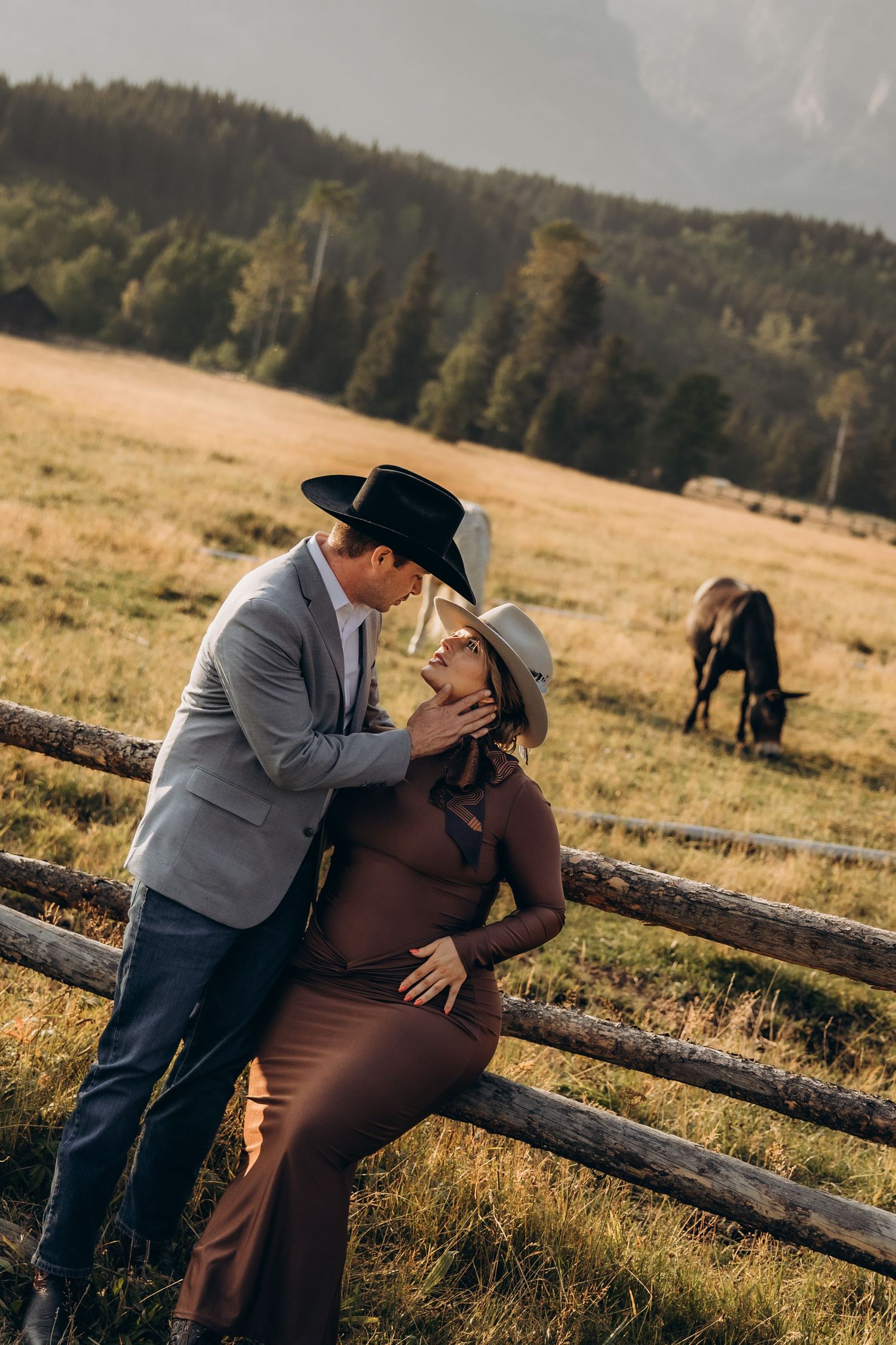 A romantic couple shares a loving moment against a rustic wooden fence at sunset in a mountain pasture.