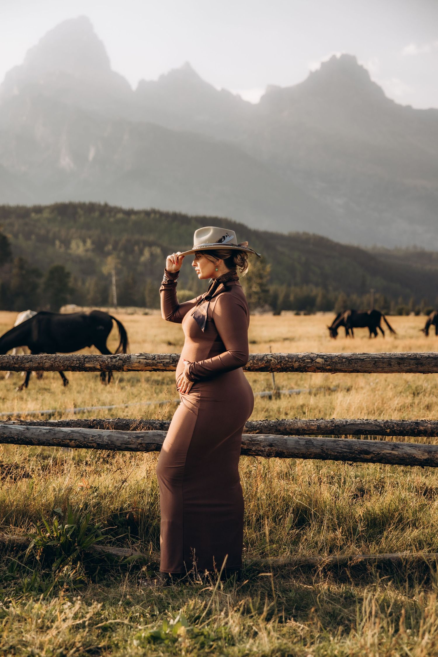 A photographer wearing brown takes photos of wild horses grazing in a golden mountain meadow during sunset.