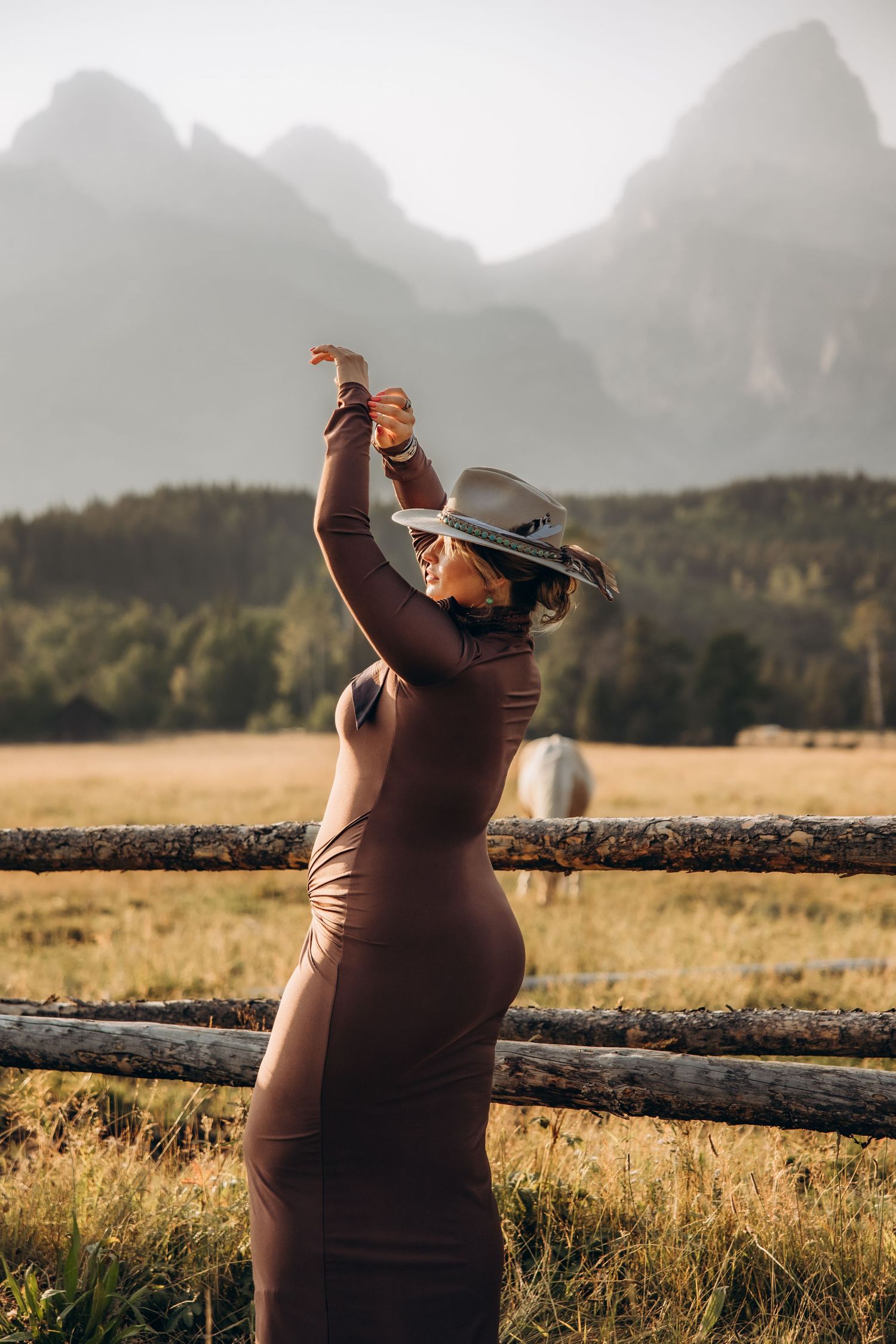 A silhouette stretches against a misty mountain backdrop at golden hour, wearing a western hat in a rustic field setting.