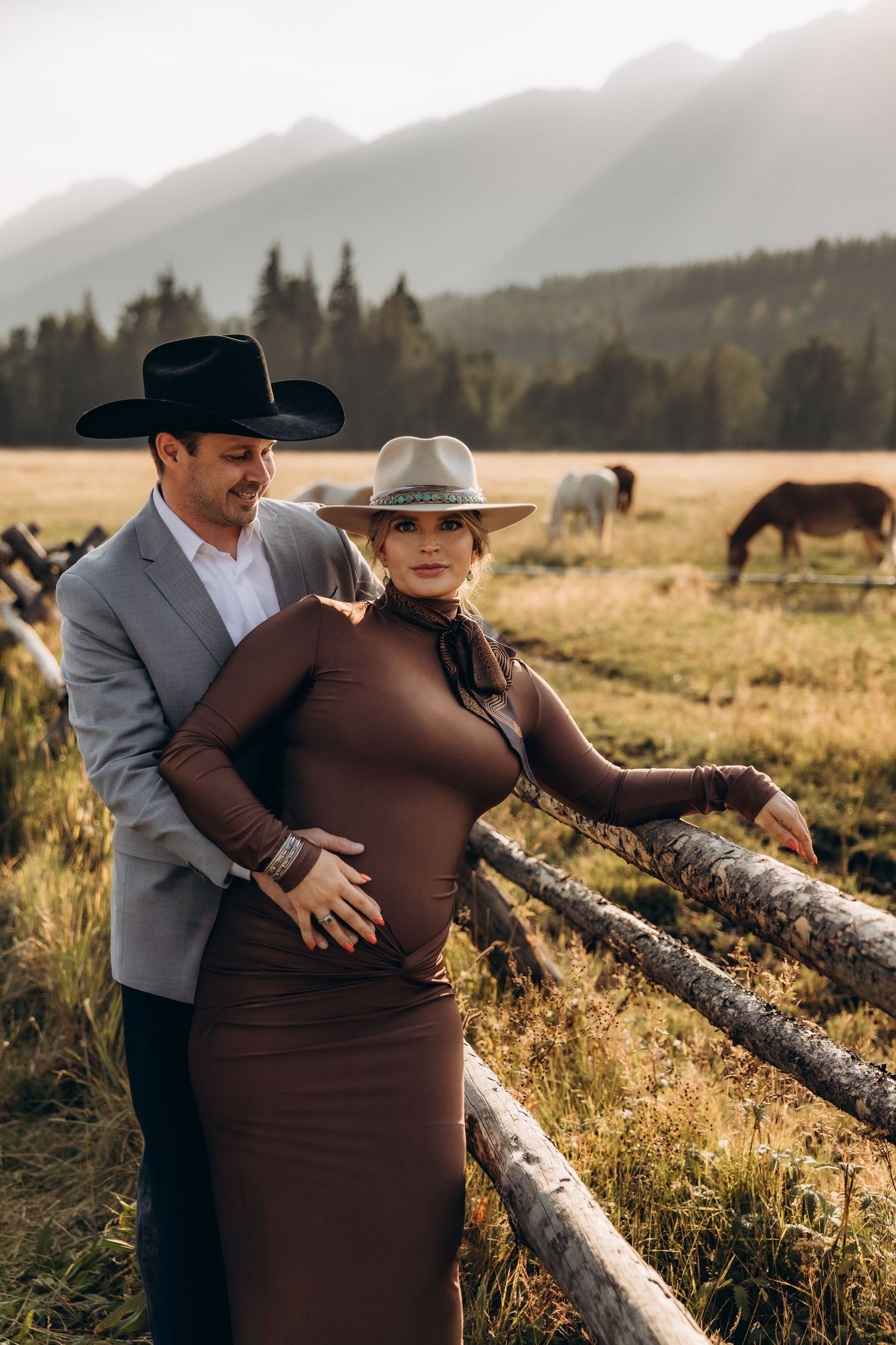 A romantic couple embracing in a rustic field at sunset with mountains and horses in the background.