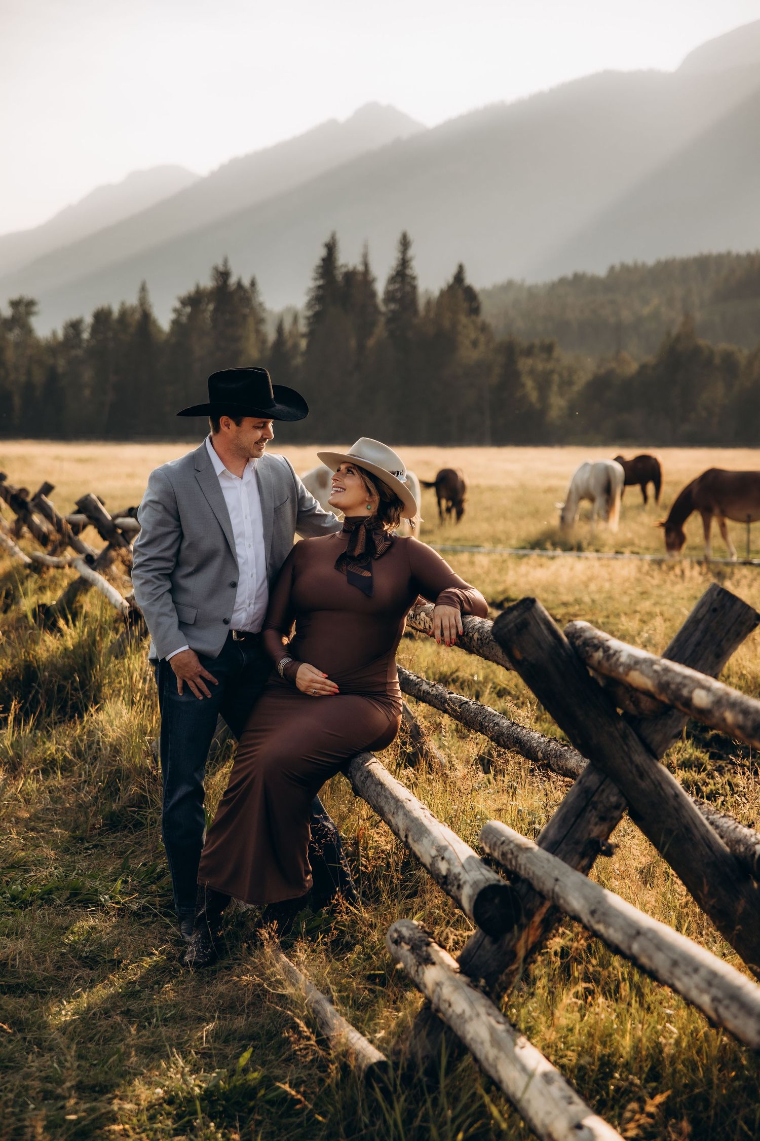 A couple sits together on rustic wooden logs in a scenic mountain meadow with horses grazing in the background.