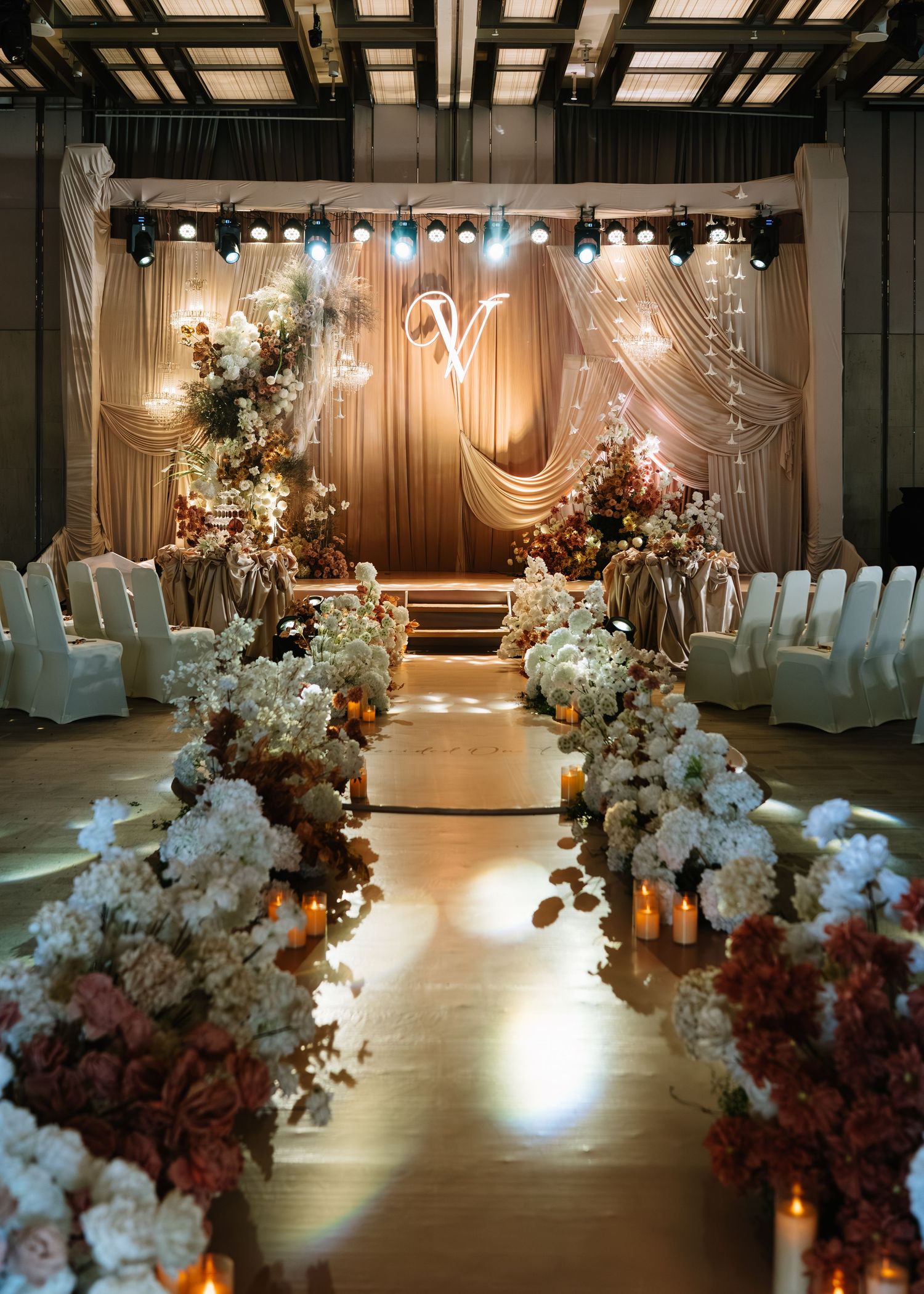 Elegant wedding ceremony aisle with white floral arrangements, candles, and a draped champagne backdrop with monogram.