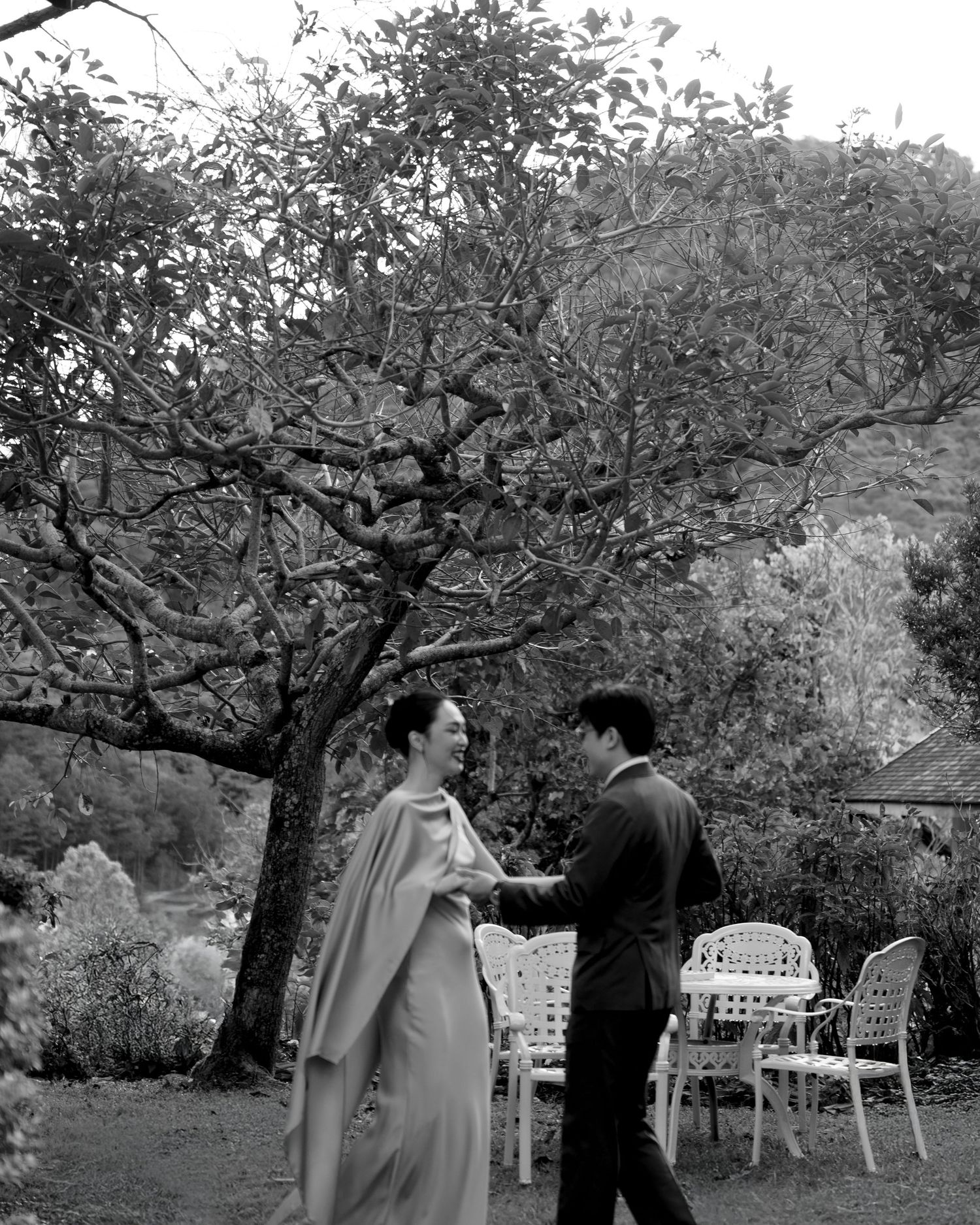 Black and white photo of a couple sharing a joyful moment in a lush garden with a large tree backdrop.