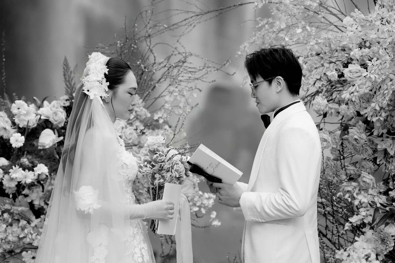Black and white wedding ceremony photo of bride and groom exchanging vows at floral altar.