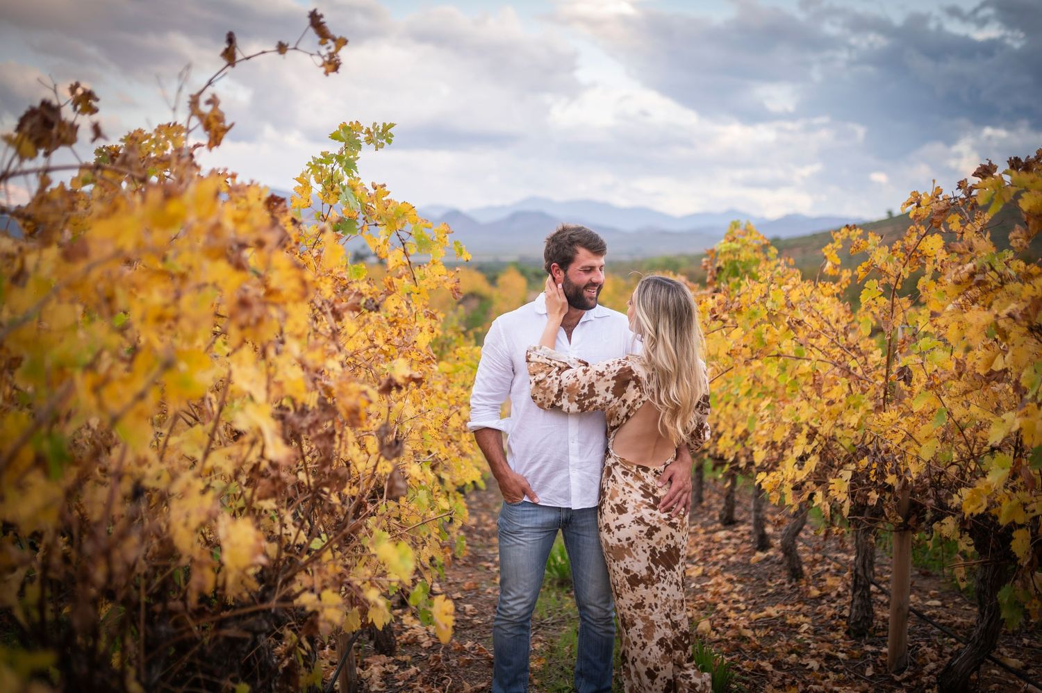 A romantic couple embraces among autumn vineyard rows with golden yellow leaves and mountains in the background.