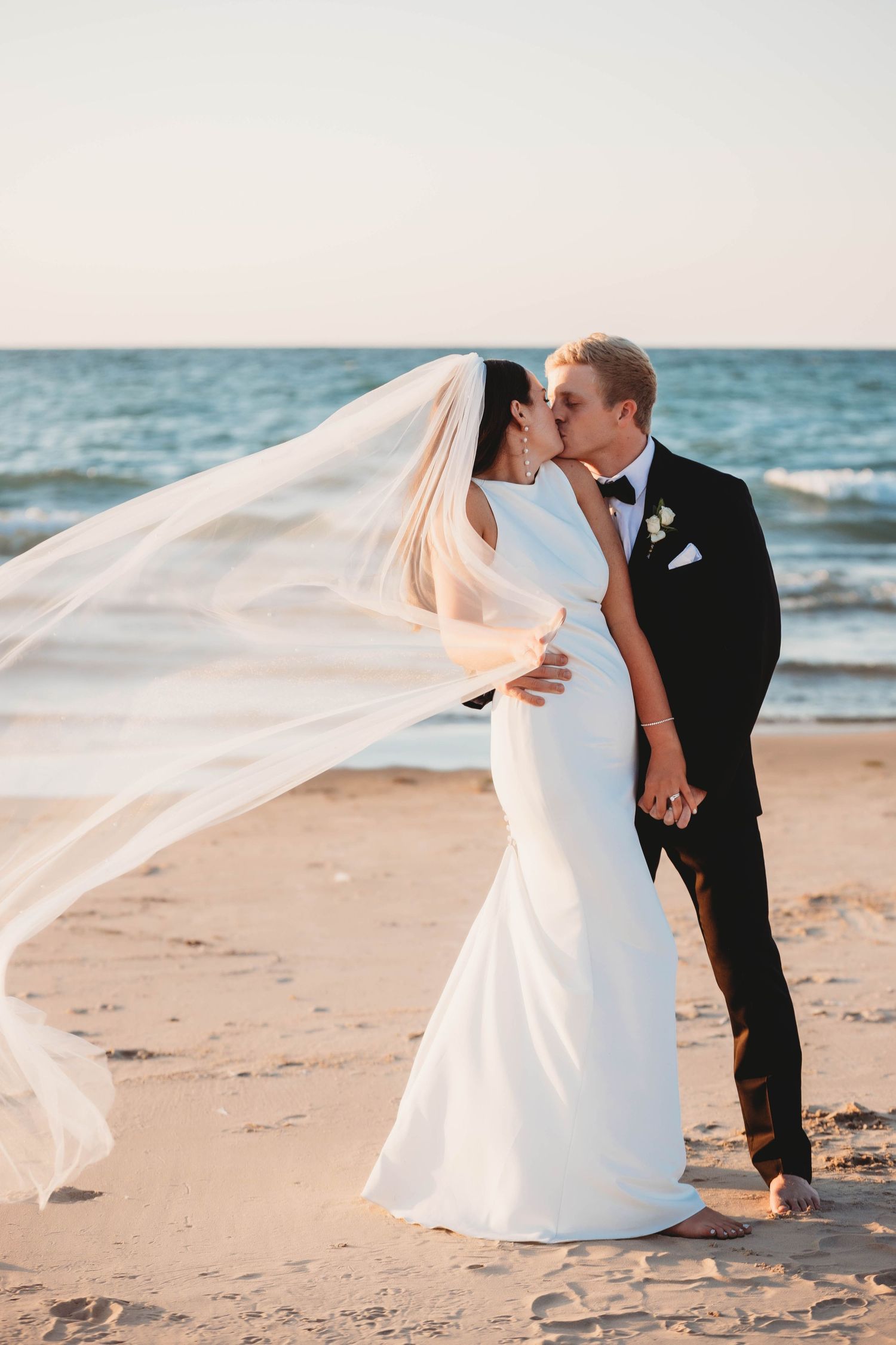 A wedding couple shares intimate moments on a beach at sunset while the bride's white veil flows in the ocean breeze.