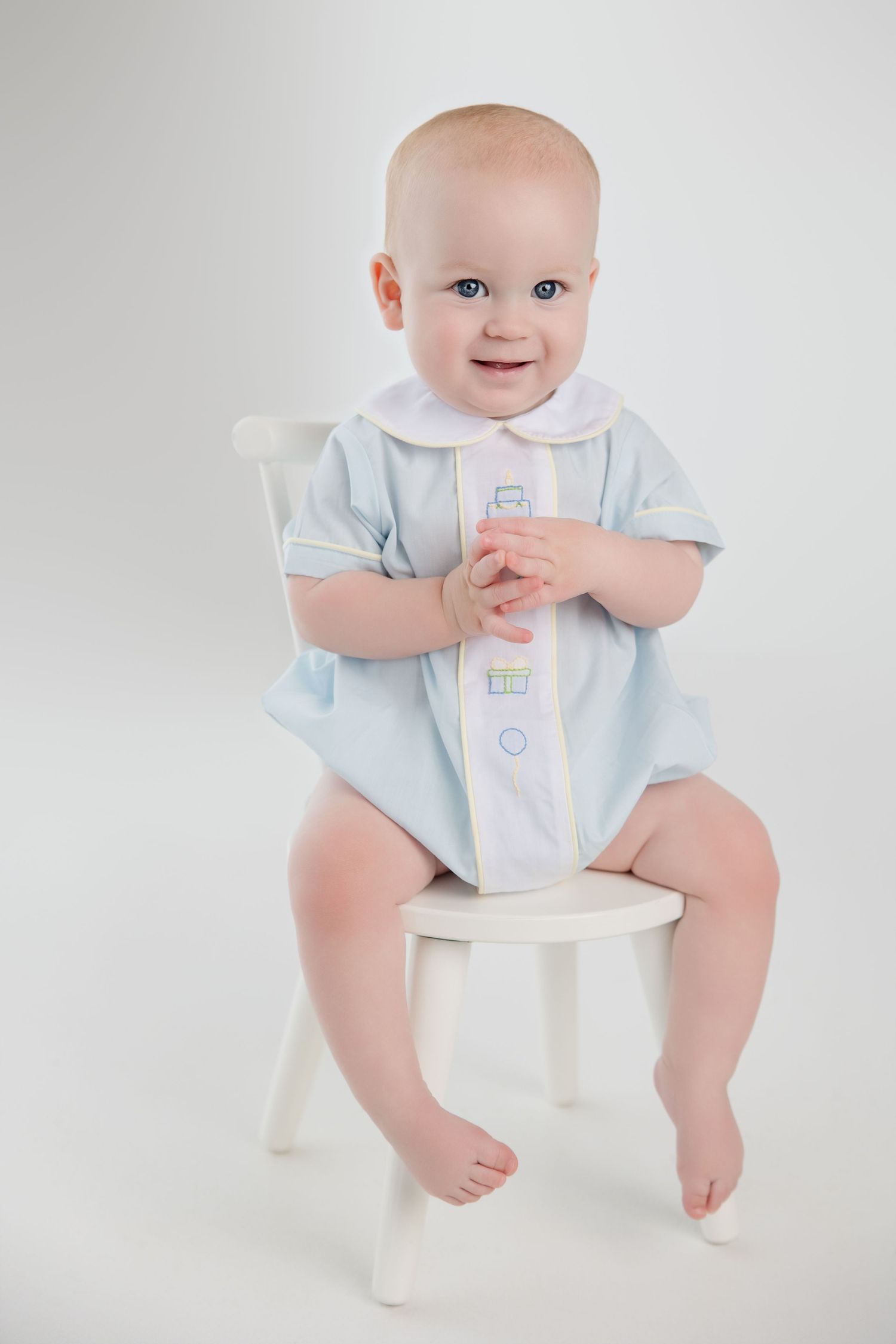 Sweet baby in light blue outfit sits on round white stool during portrait session.