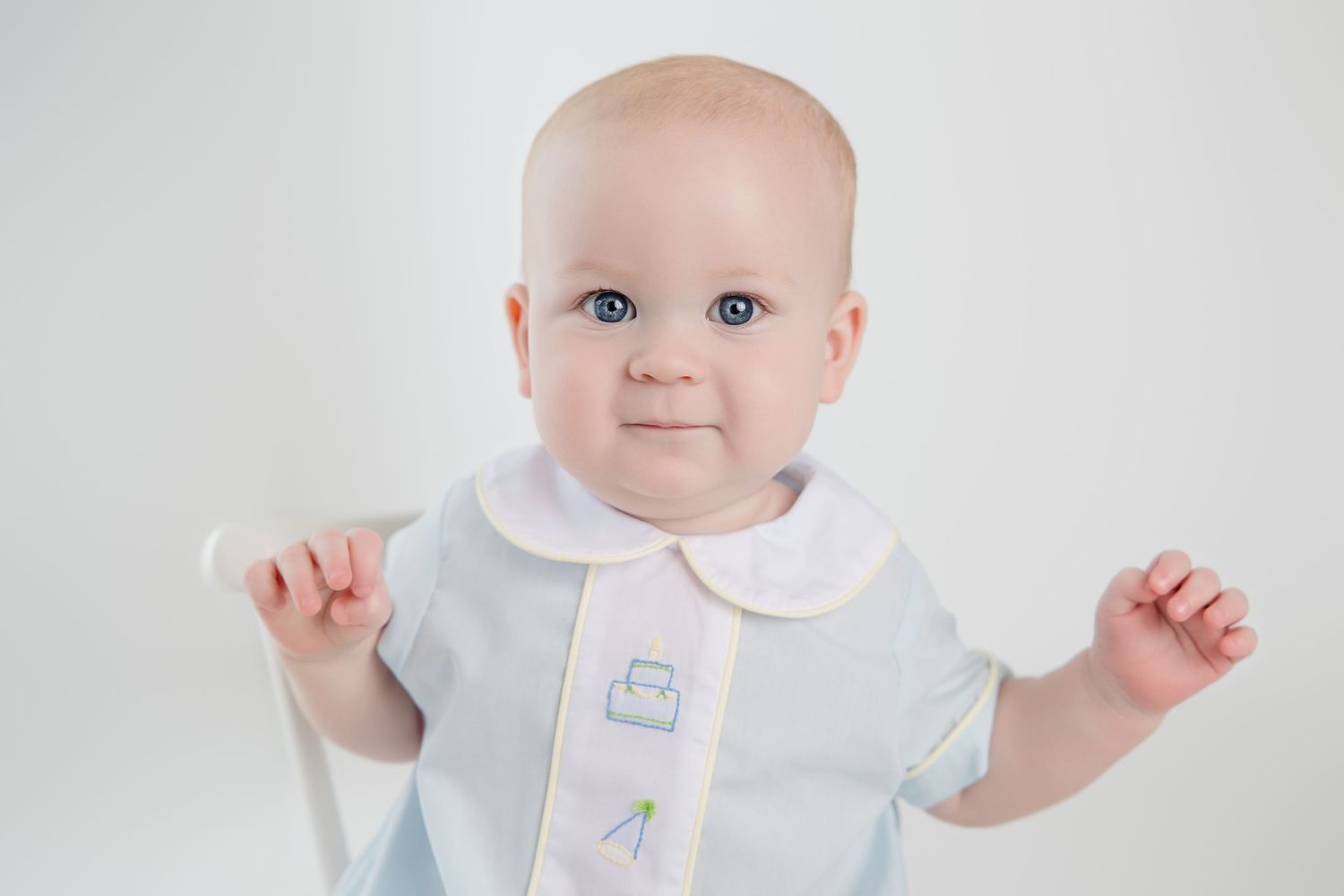A baby wearing white clothing looks at the camera with bright blue eyes against a white background.