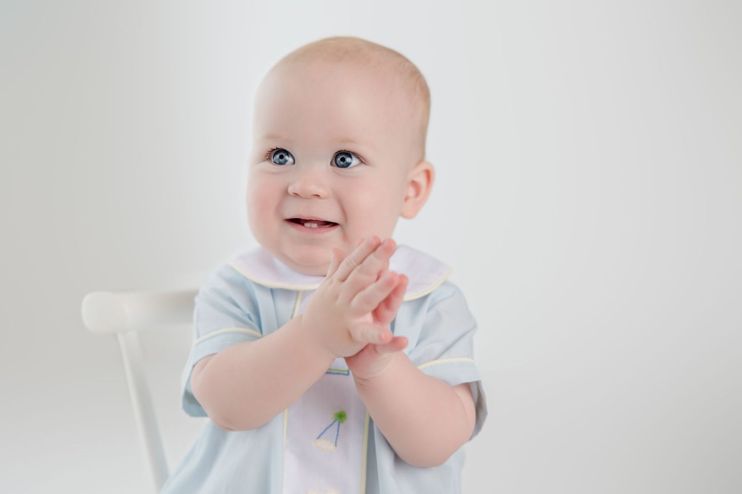 A happy baby in light blue clothing sits and smiles brightly while covering their mouth with one hand.