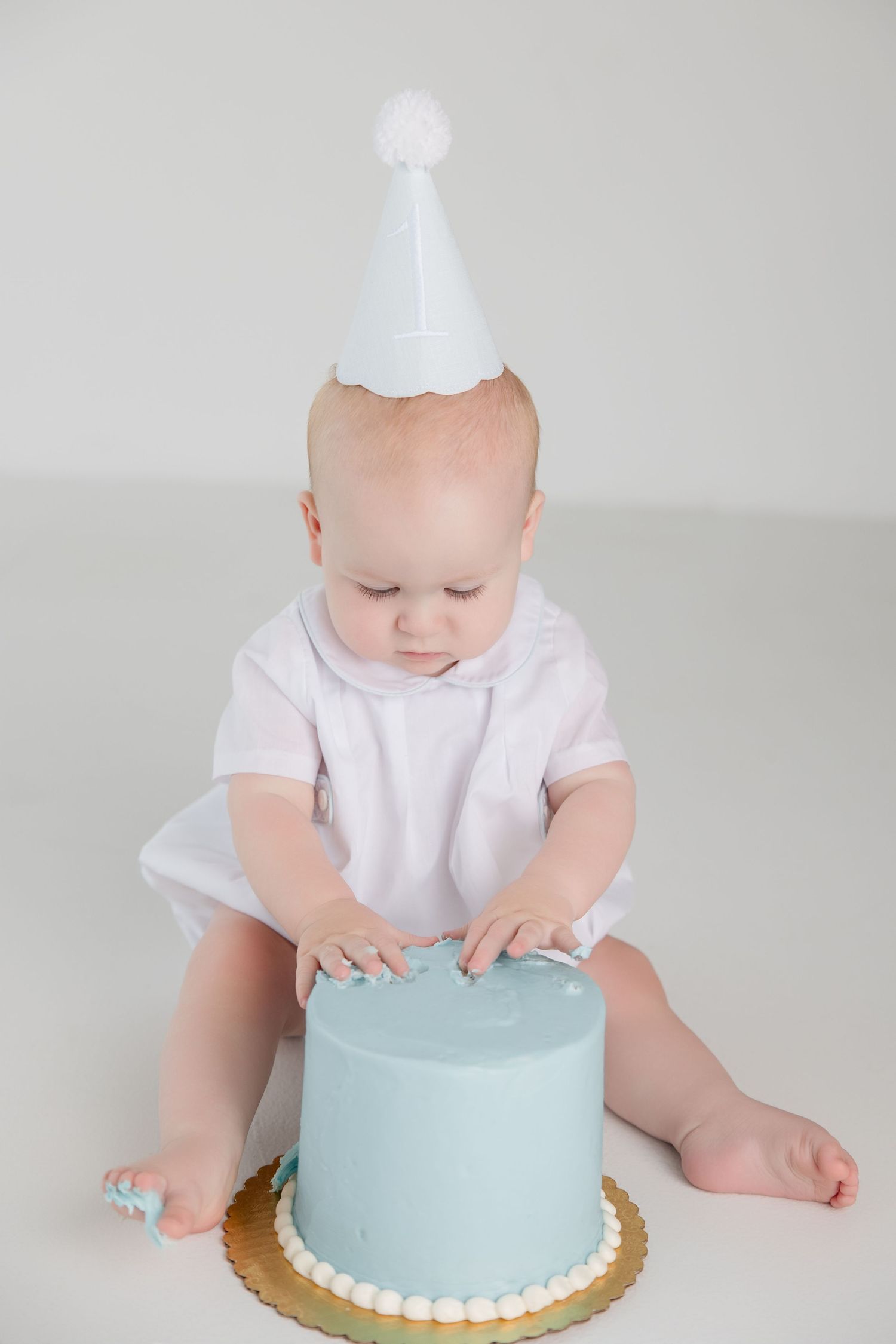 Baby in white outfit exploring a light blue birthday cake on a gold base against grey background.