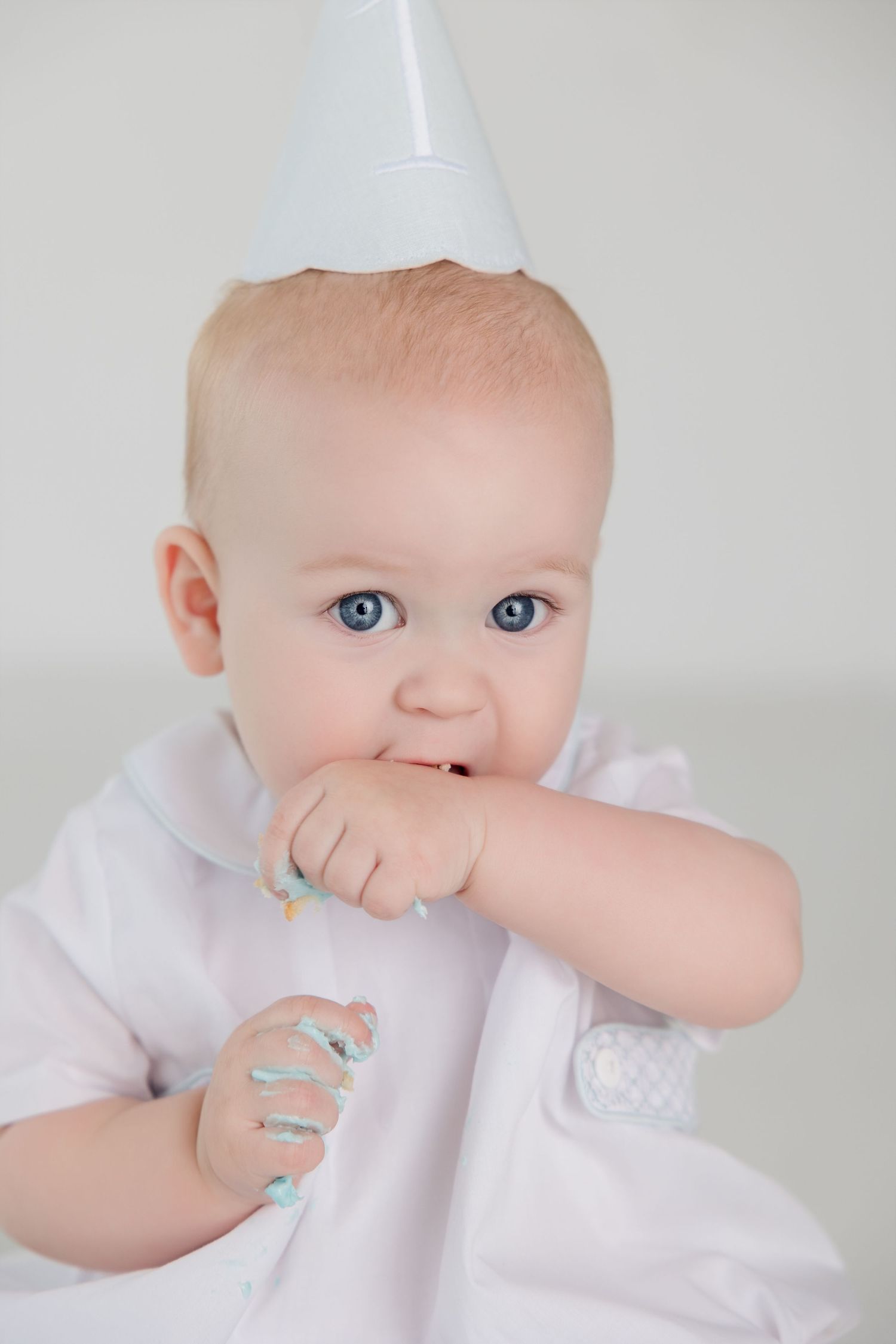 Baby wearing white outfit getting messy while eating blue frosted cake.