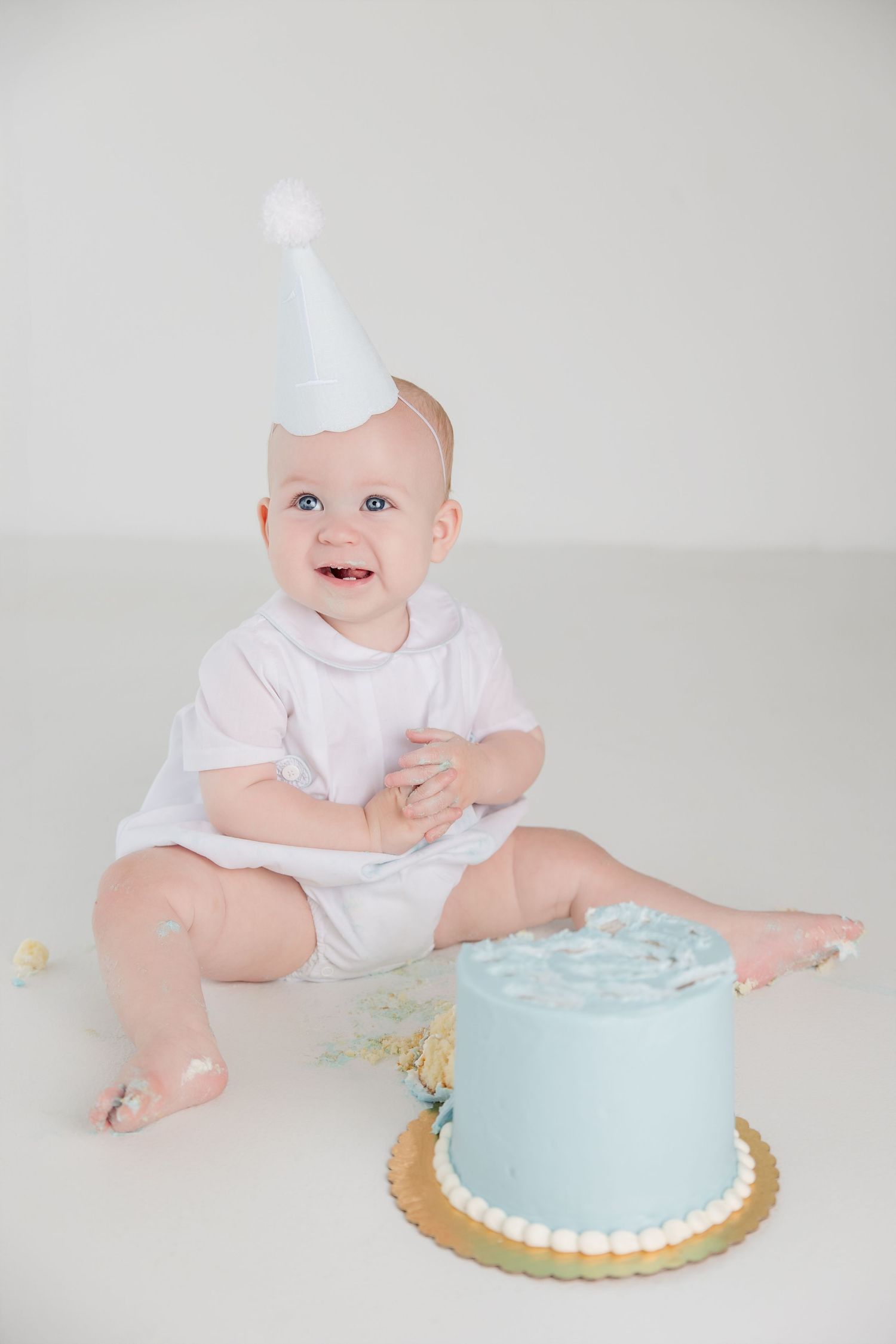 Baby in white outfit celebrates first birthday with light blue smash cake in minimalist studio setting.