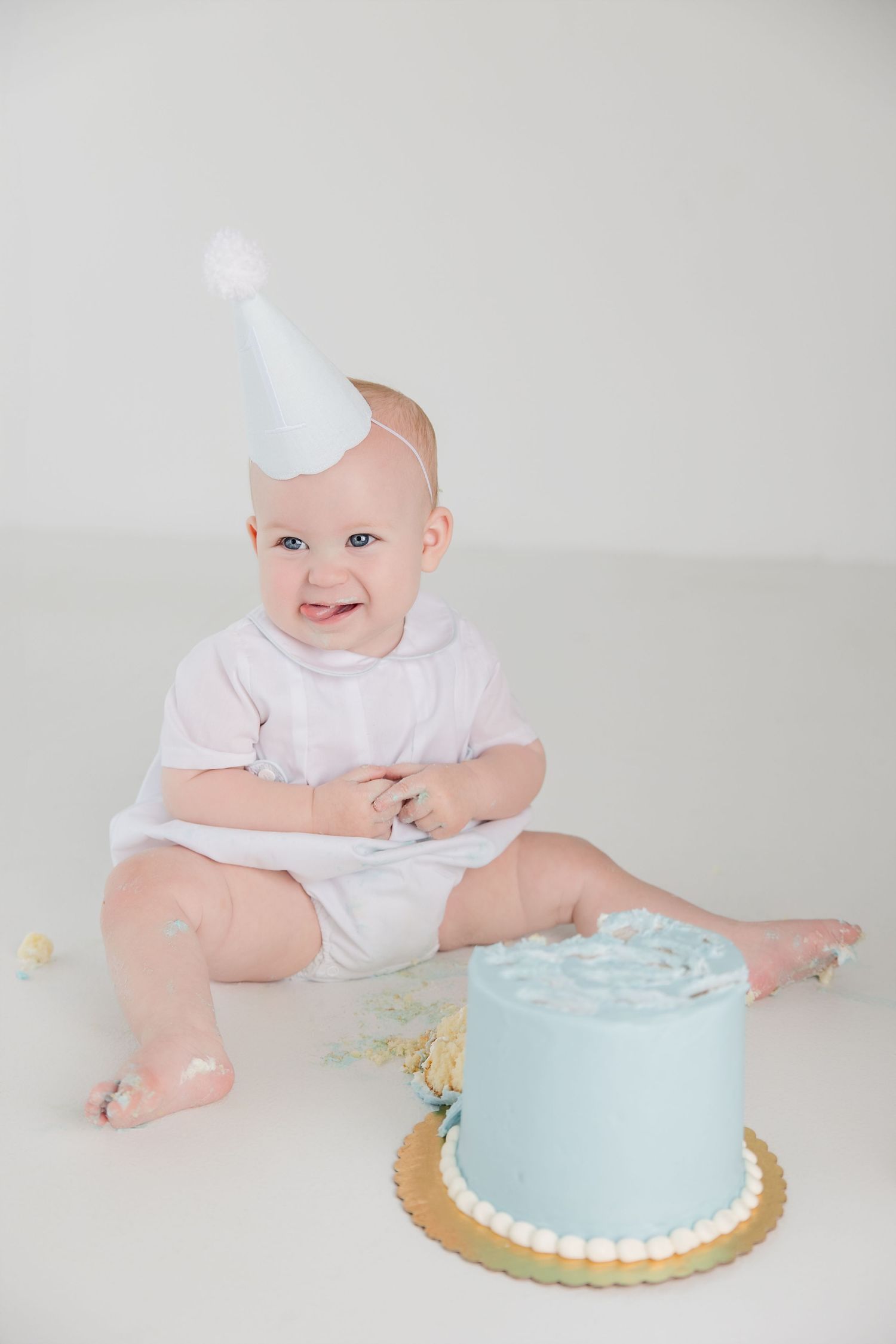 Baby in white outfit sitting and smiling next to light blue cake with white trim.