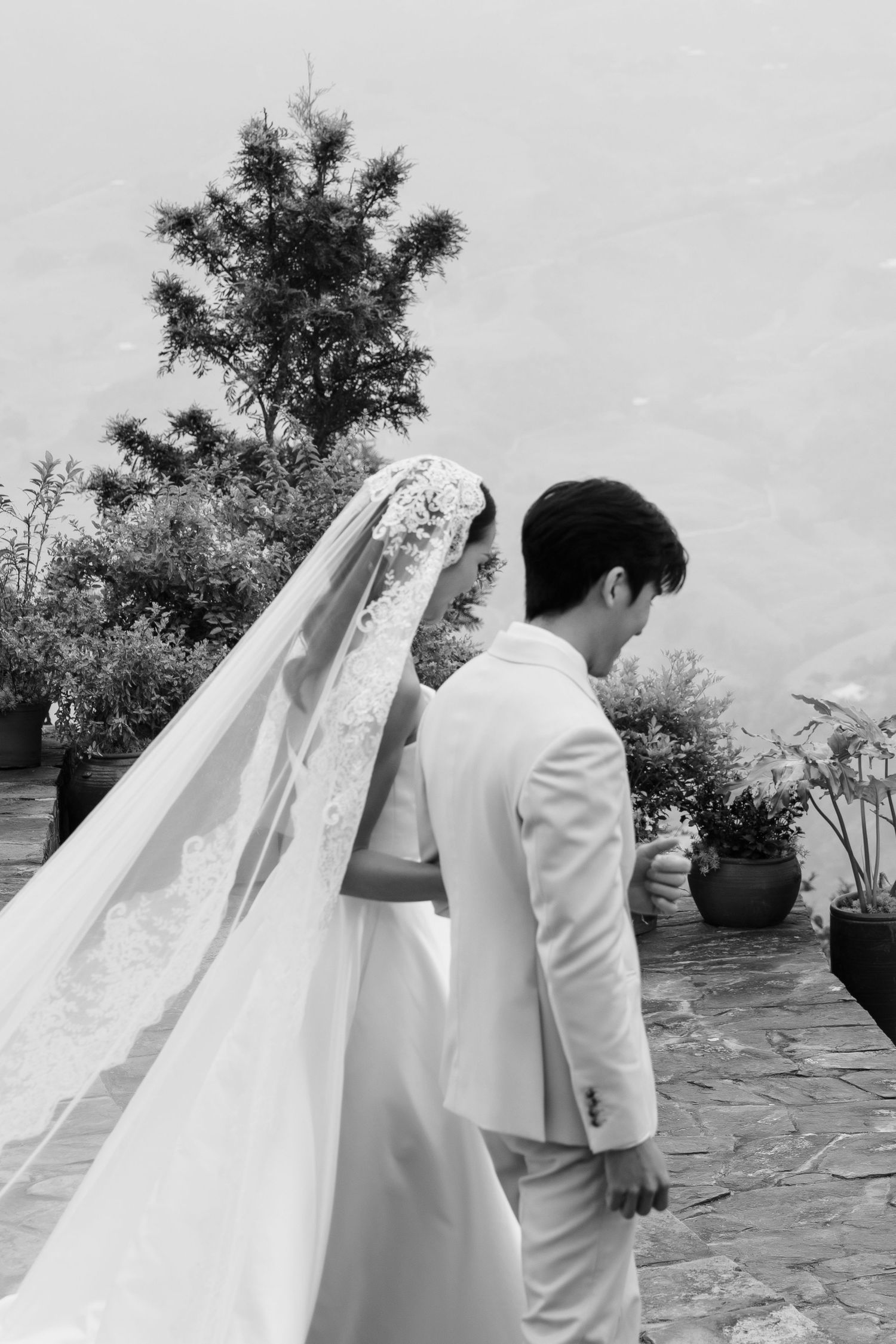 Black and white portrait of a bride in a lace veil and groom in a white suit walking together among lush greenery.
