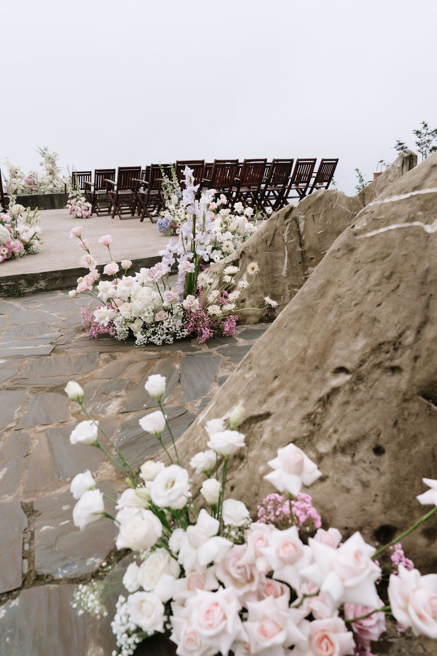 Close-up of pink and white wedding floral arrangements along a stone aisle with wooden chairs.