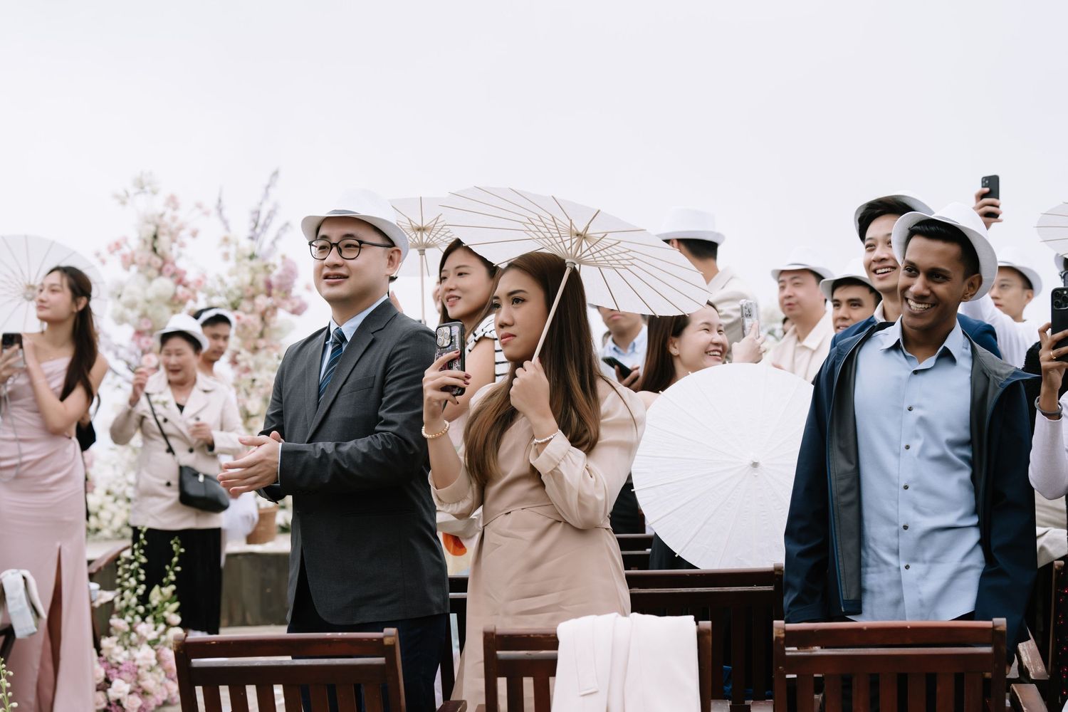 Wedding guests smile and hold white parasols during an outdoor ceremony with pink floral arrangements in the background.
