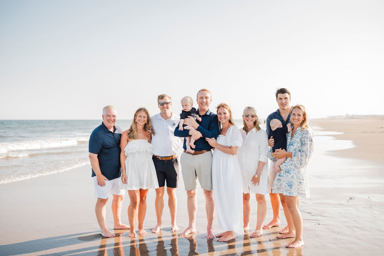 A group relaxes together on a sunny beach wearing coordinated white and blue outfits for photos at sunset.