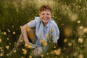 Child in a blue shirt smiles while sitting in a golden summer field at sunset.