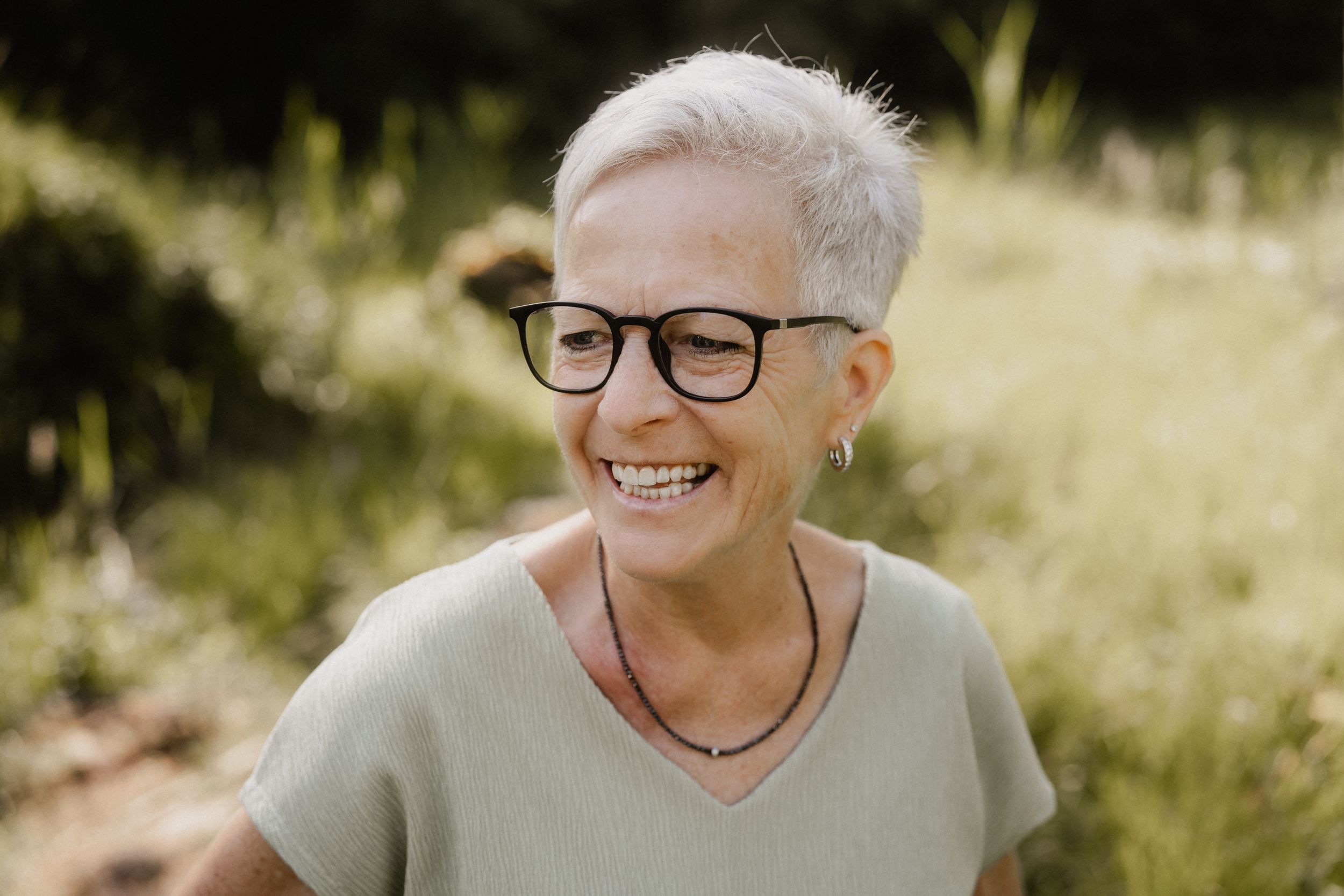 A person in glasses smiles joyfully while sitting in a sunlit meadow with tall grass and trees in the background.