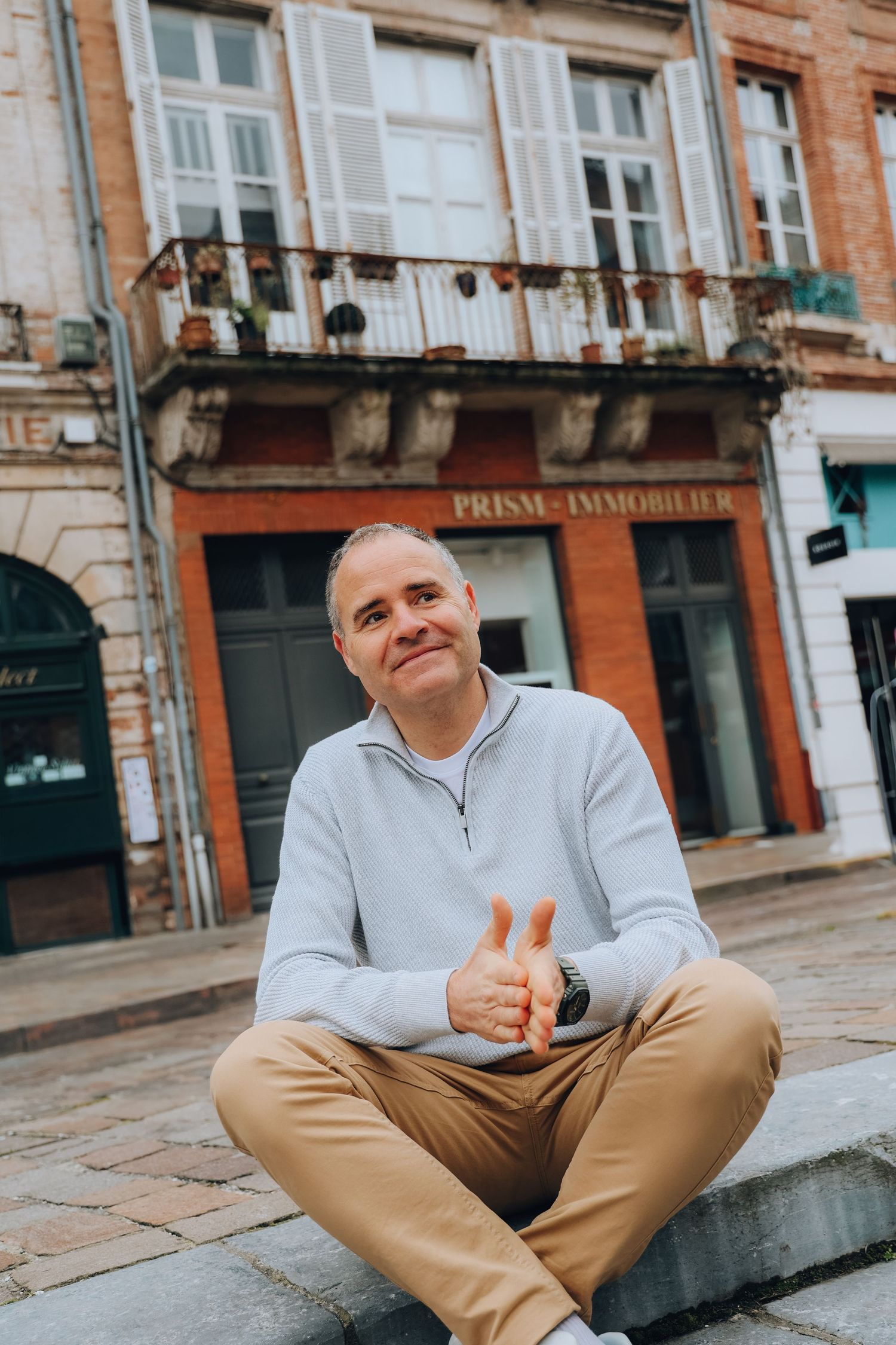 A cheerful man in a light blue sweater and khaki pants sits cross-legged giving thumbs up in a European town square.