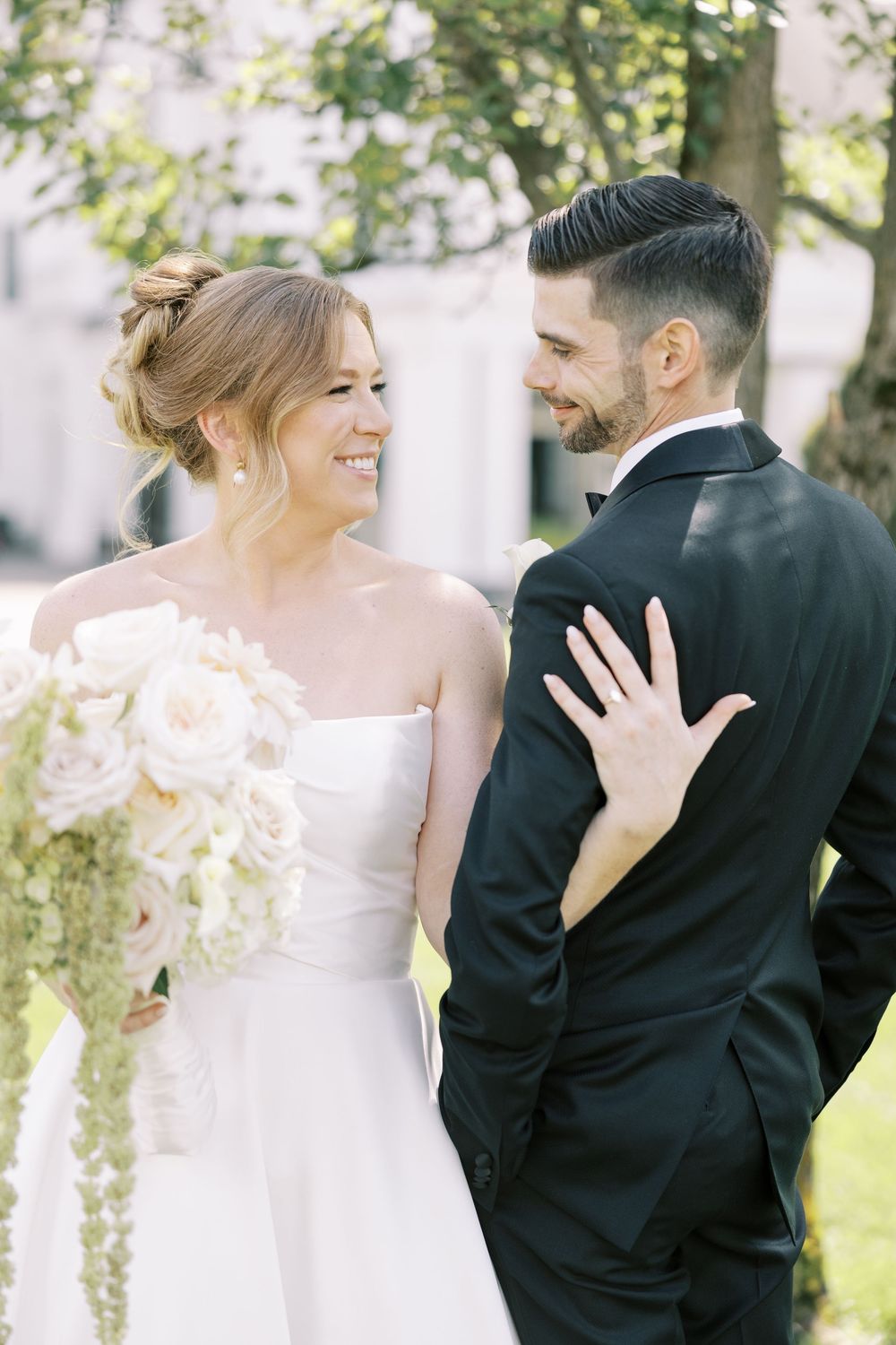 A couple in wedding attire share a joyful moment in a sunny garden setting with blush pink bouquet details.