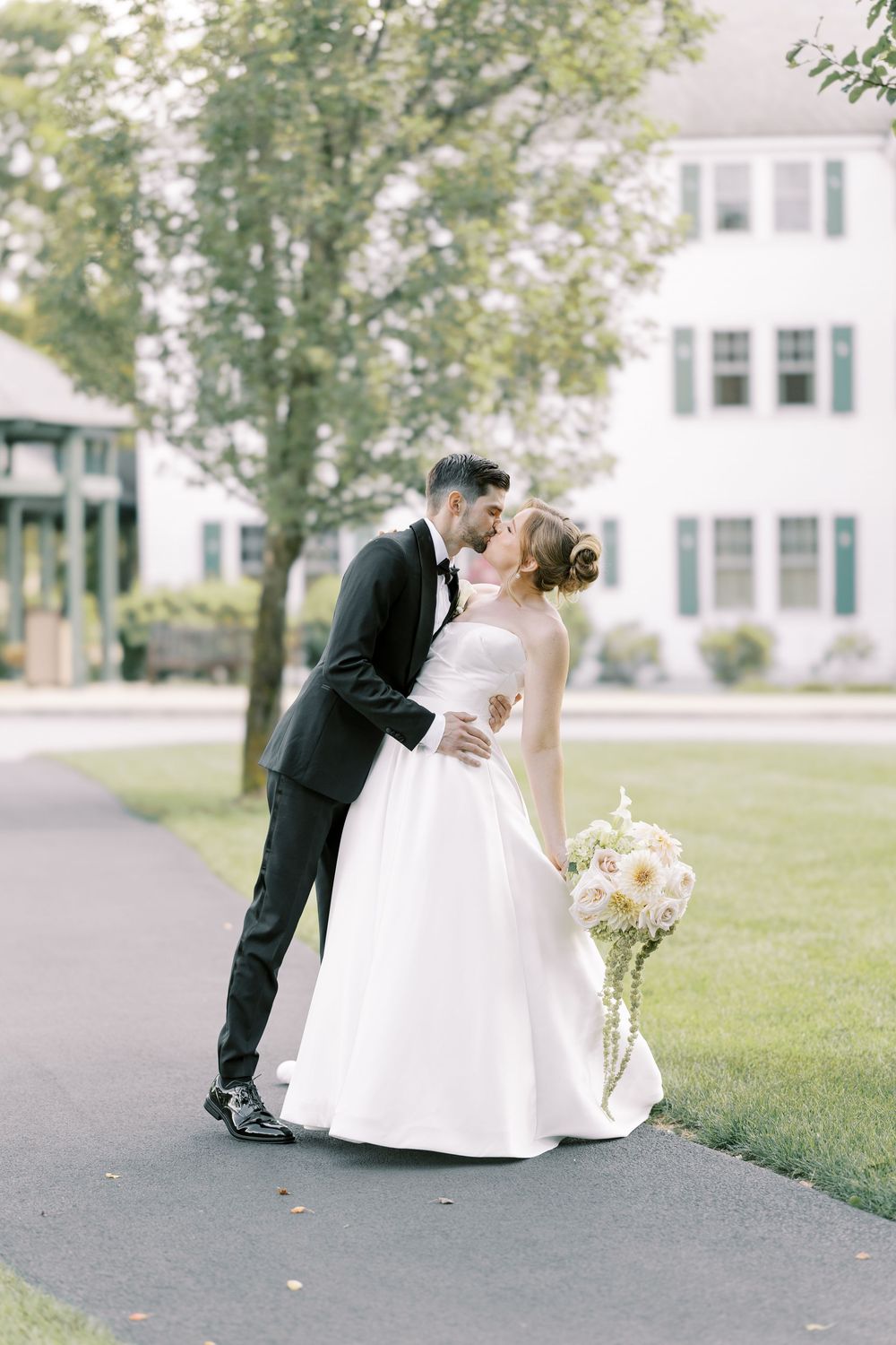 A romantic wedding couple shares an intimate moment on a paved path with white buildings and trees in the background.